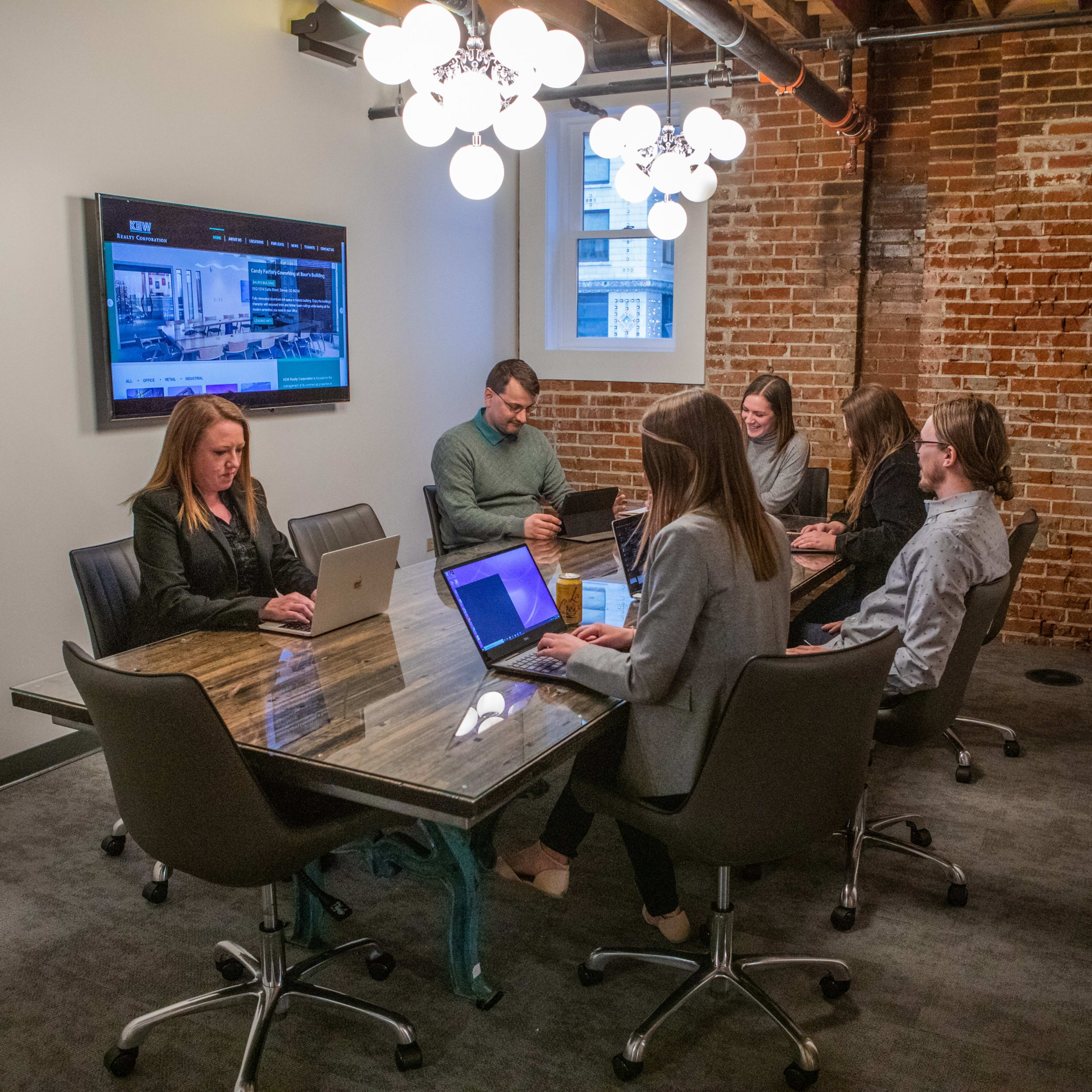 A group of six people, seated around a conference table, works on laptops in a meeting room with exposed brick walls and modern lighting.