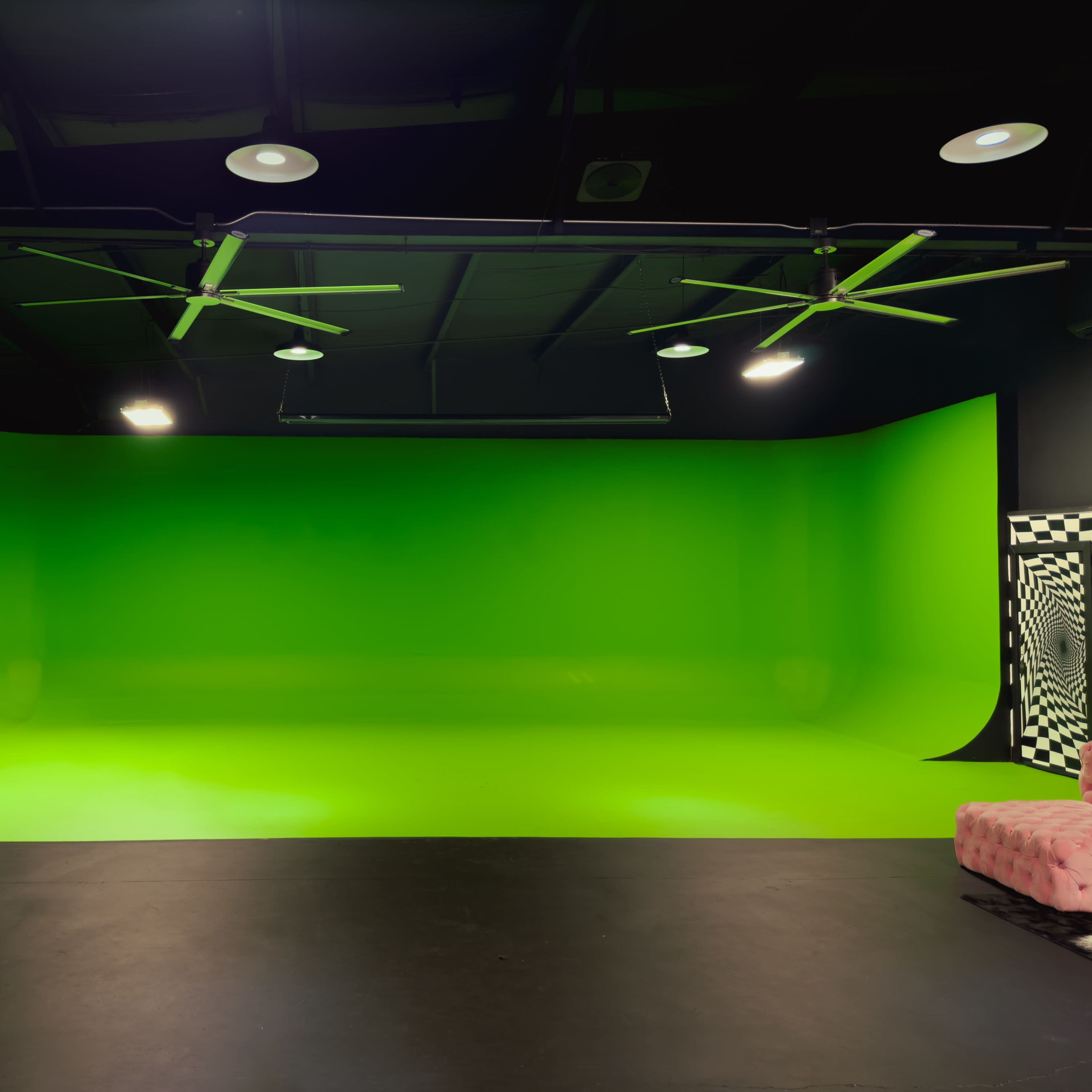 A studio with a bright green backdrop and a black floor, illuminated by ceiling lights and fans.