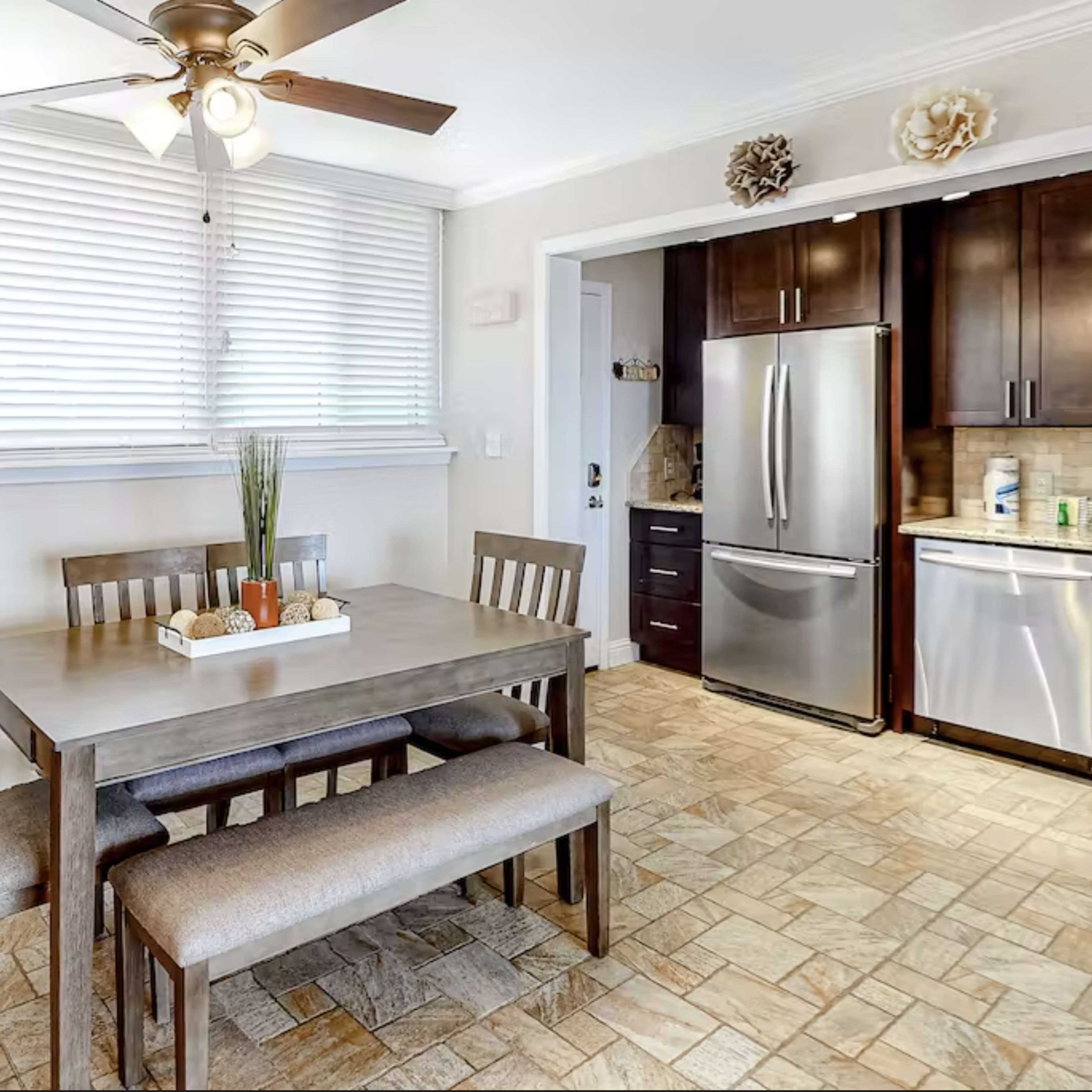 A dining area features a wooden table with benches, surrounded by light-colored walls and a kitchen with stainless steel appliances in the background.