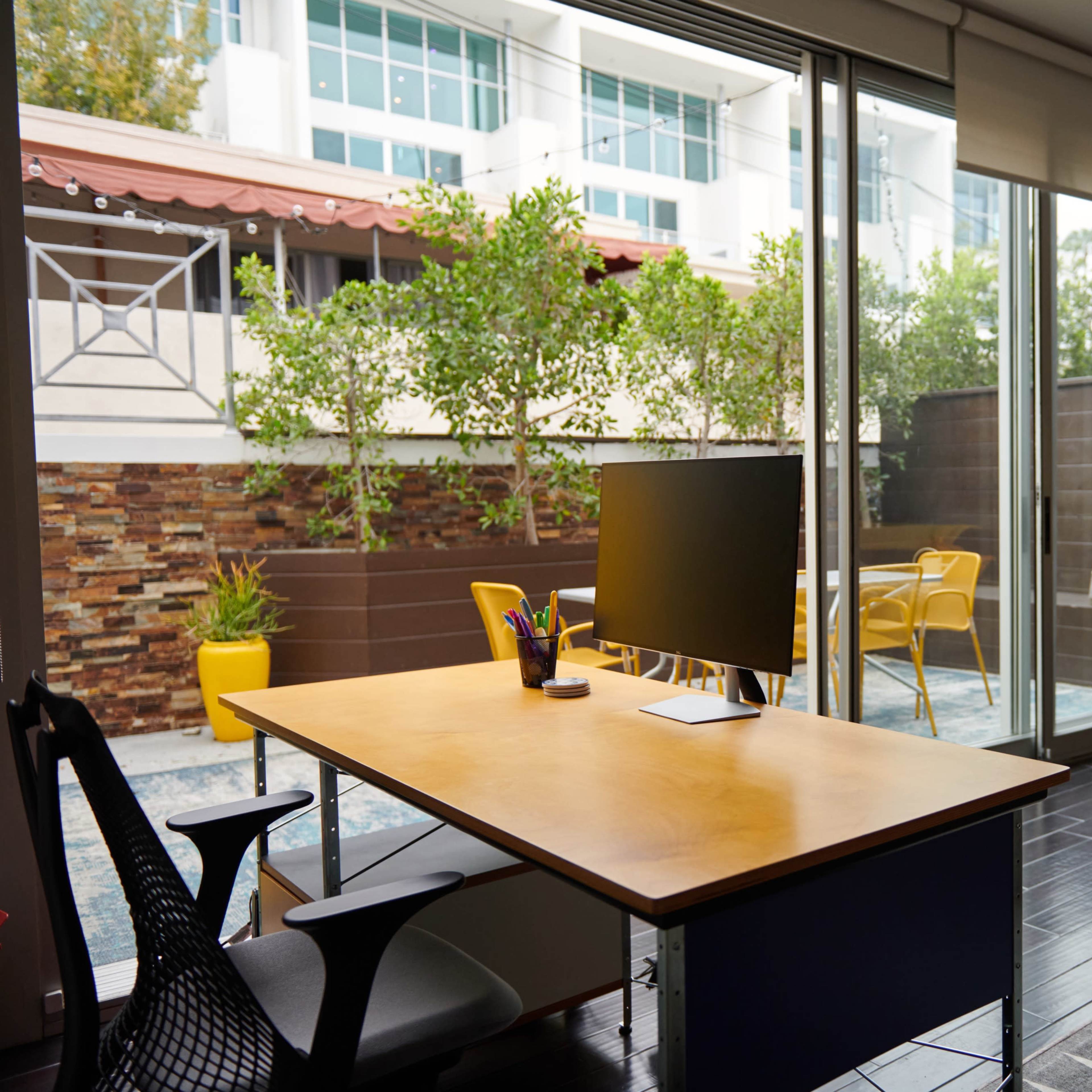A home office features a desk with a monitor, overlooking a patio with plants and yellow chairs through large glass doors.