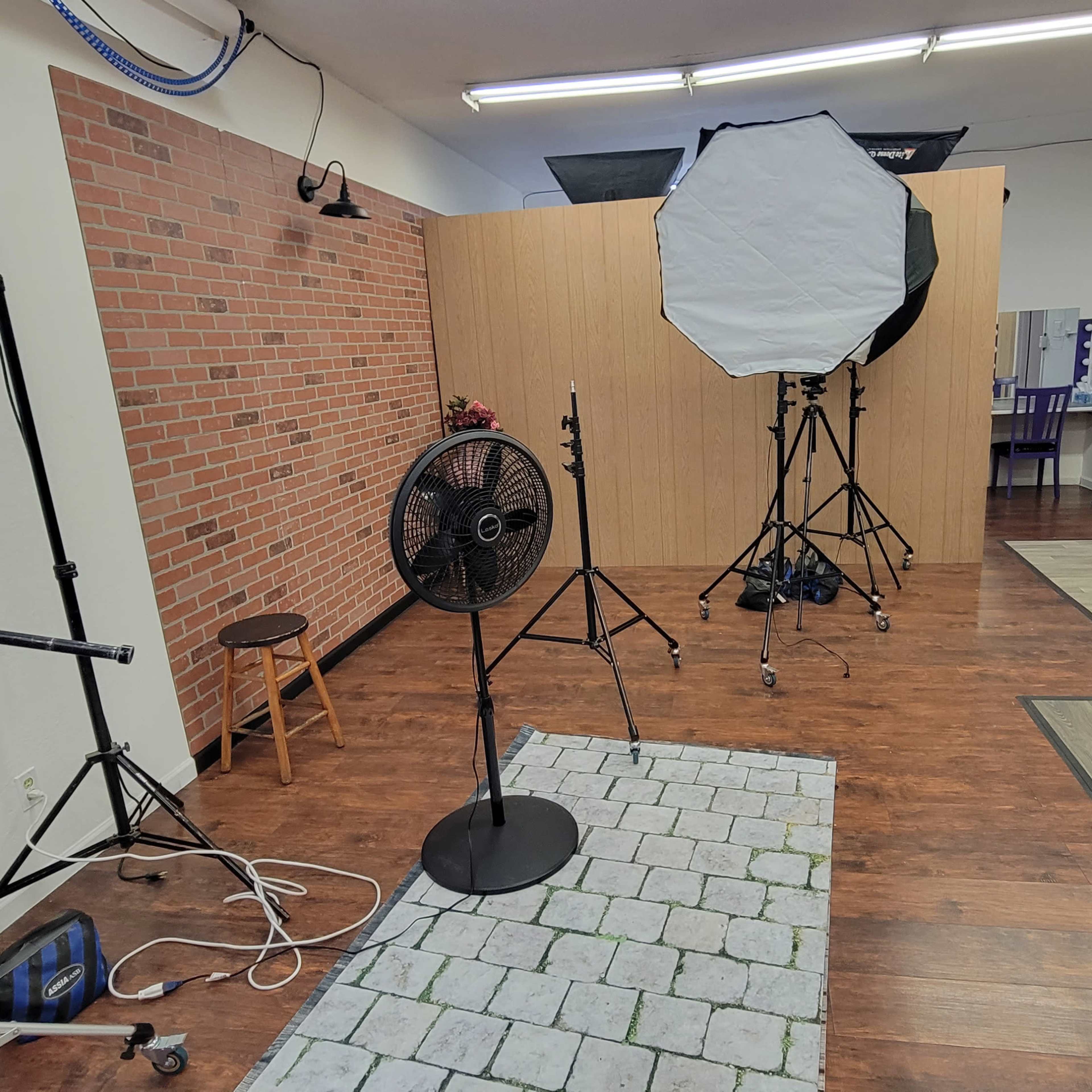 A photography studio with a brick wall backdrop, lighting equipment, a fan, and various types of furniture scattered around.