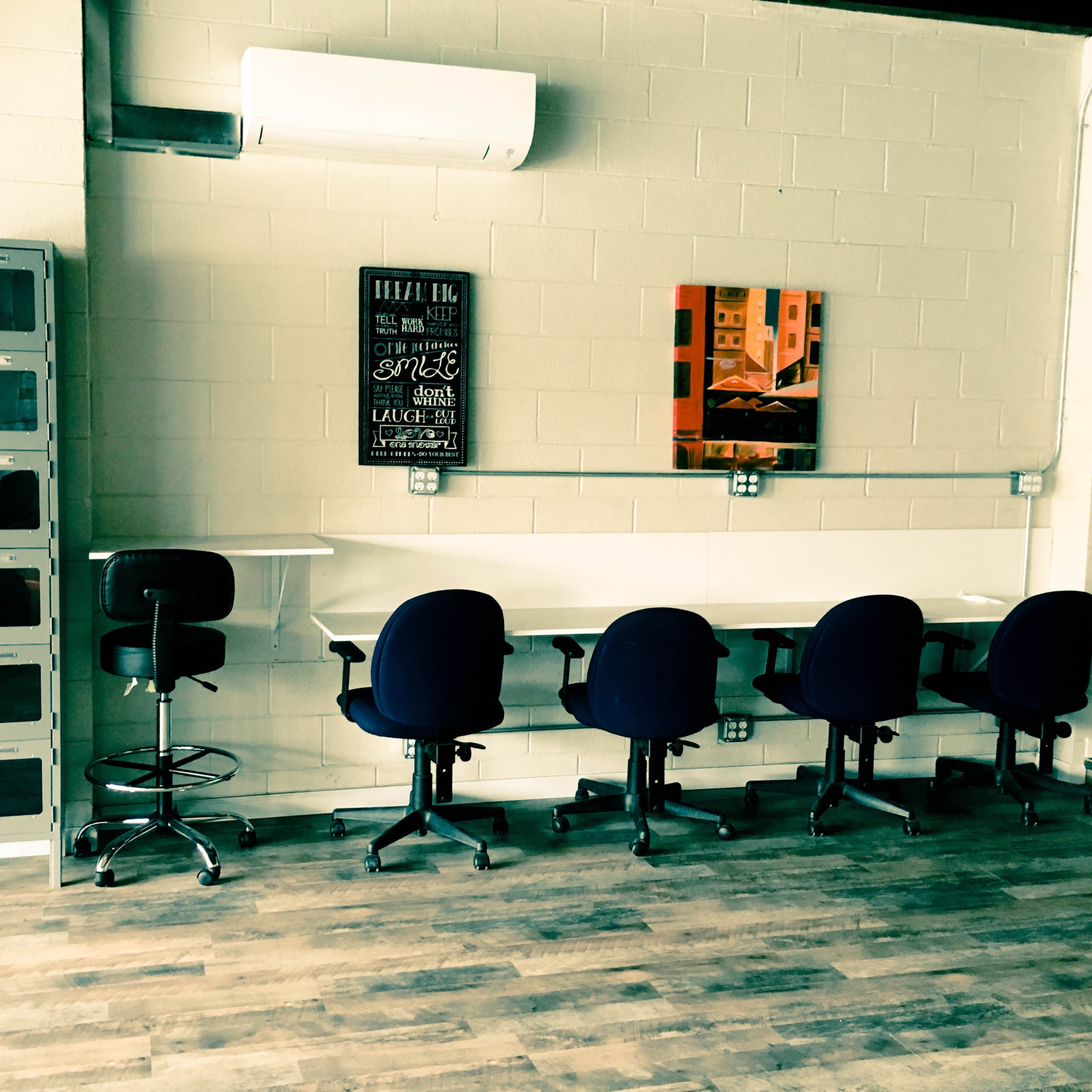 A workspace with several office chairs arranged around white desks against a light-colored wall, accompanied by a piece of artwork and a set of lockers.