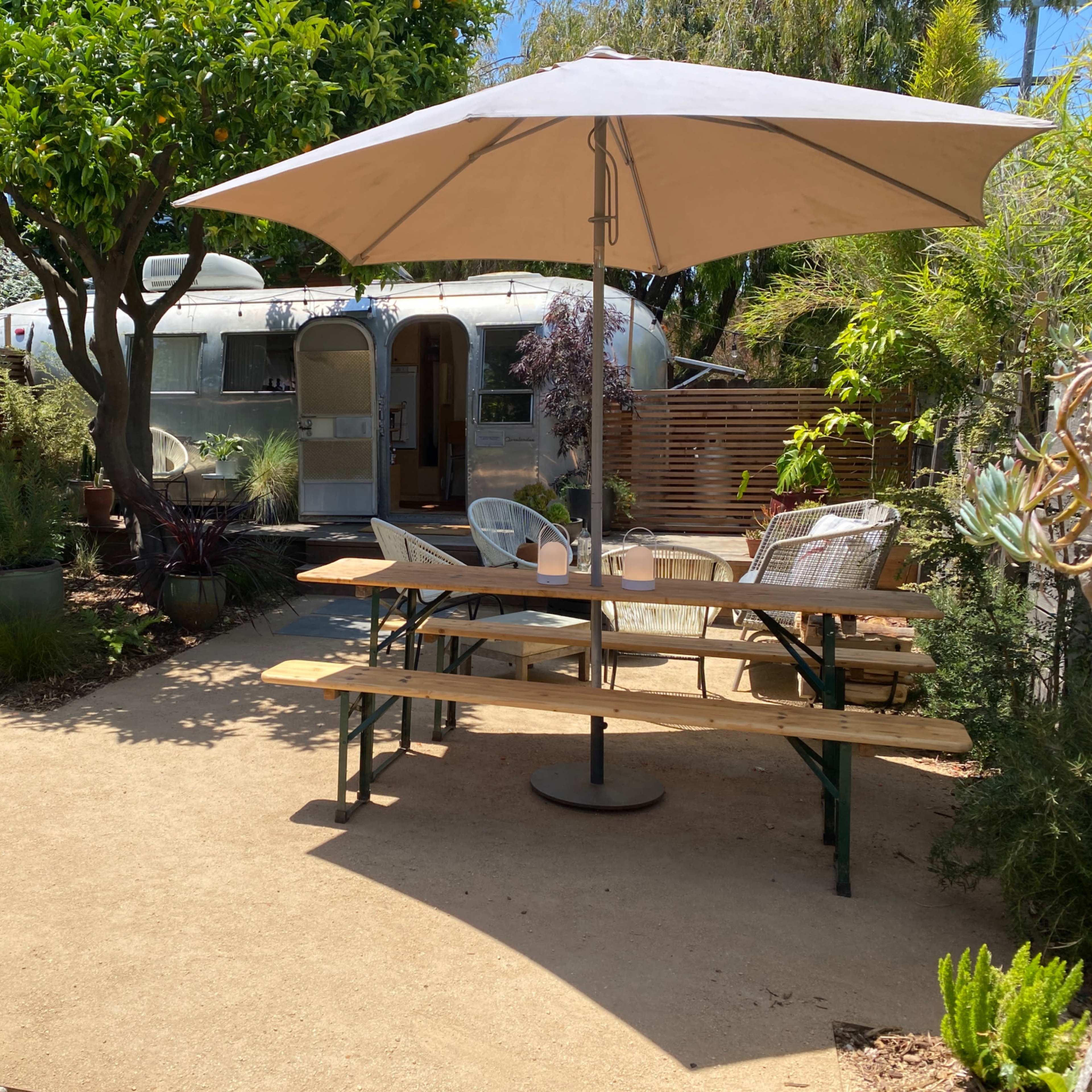 A backyard patio features a wooden table under a large umbrella, surrounded by potted plants and a vintage trailer in the background.