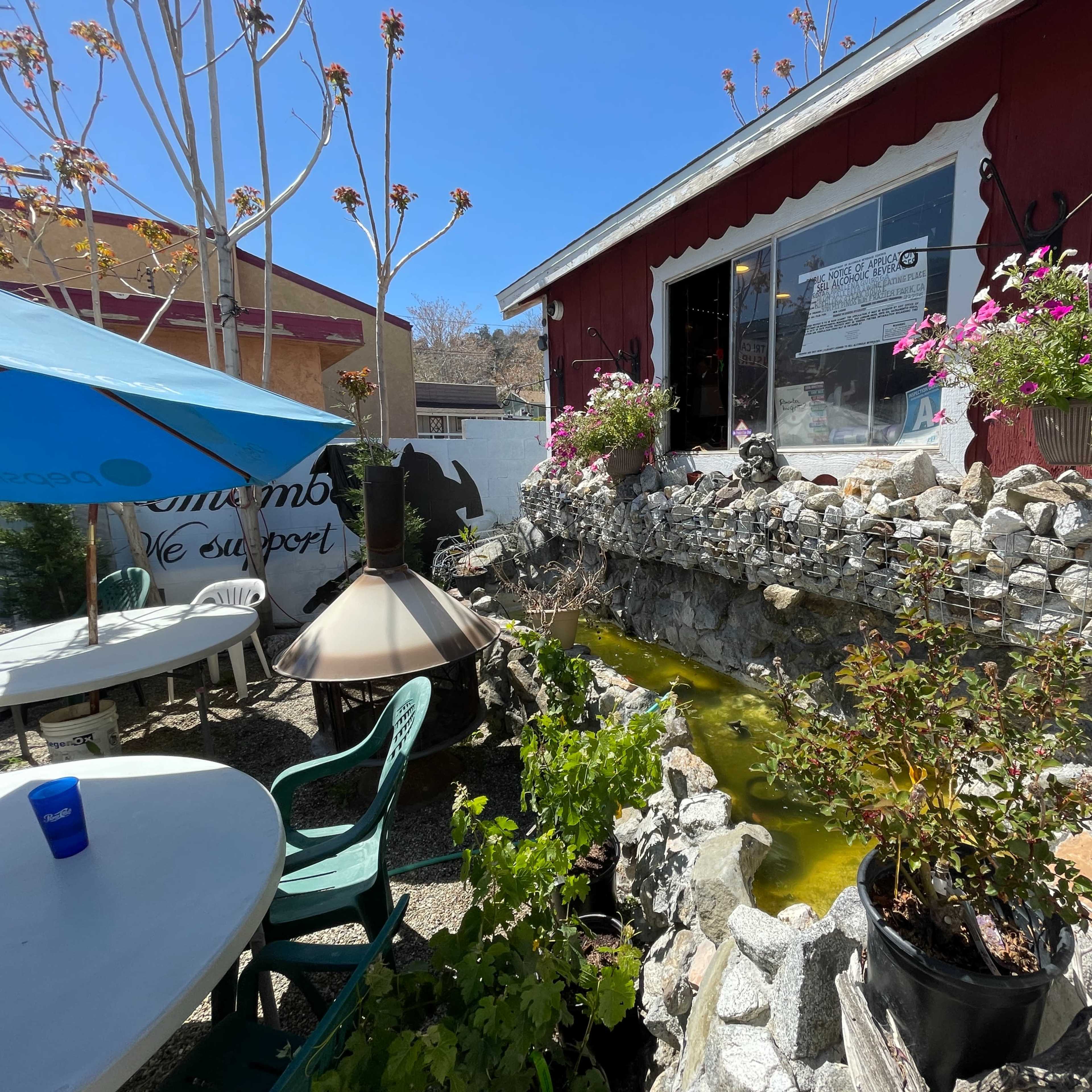 A stone-lined pond with plants and flowers is situated beside a red building with a window, outdoor tables, and a blue umbrella.
