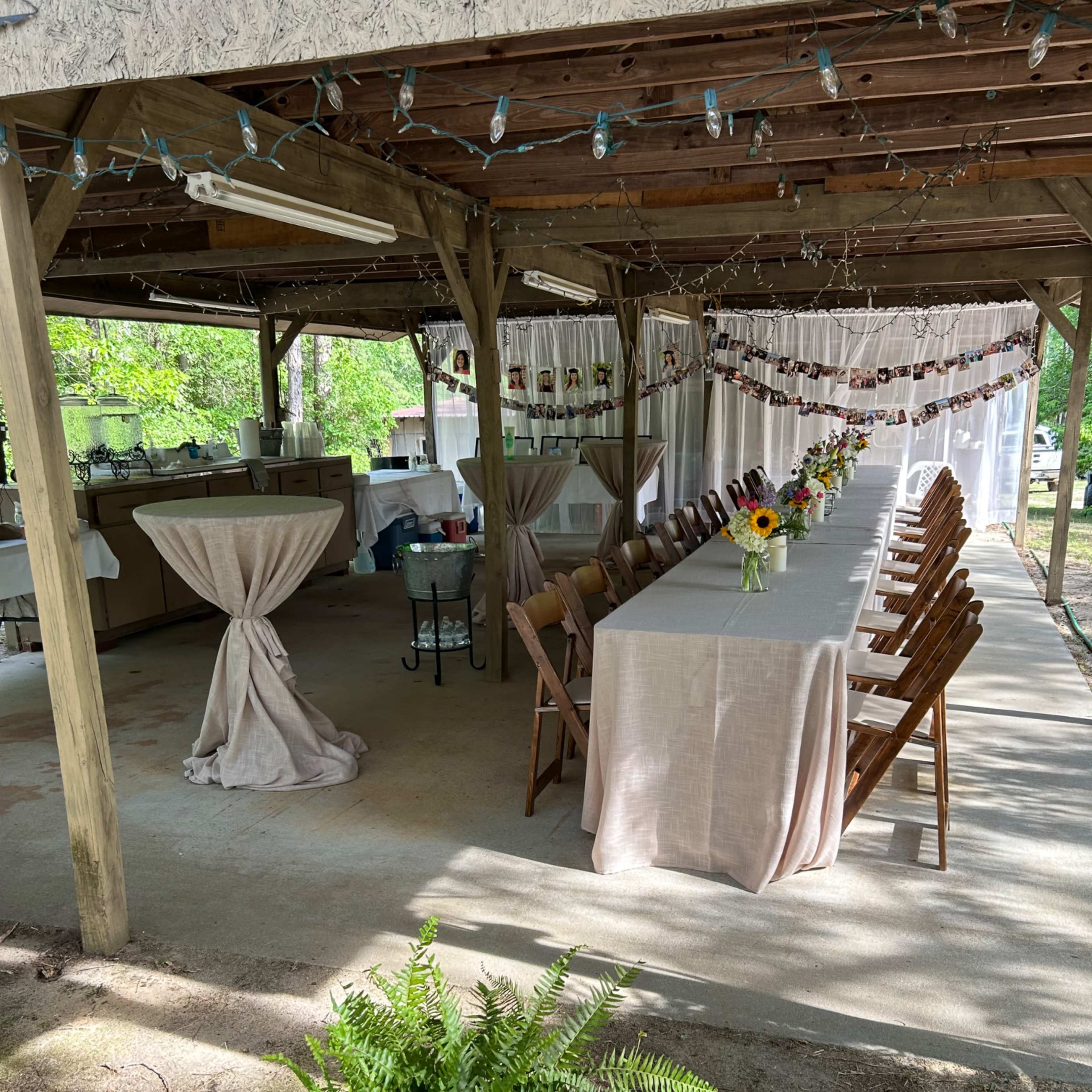 A covered outdoor space with a long table set for a gathering, surrounded by chairs and decorated with lights and flowers.