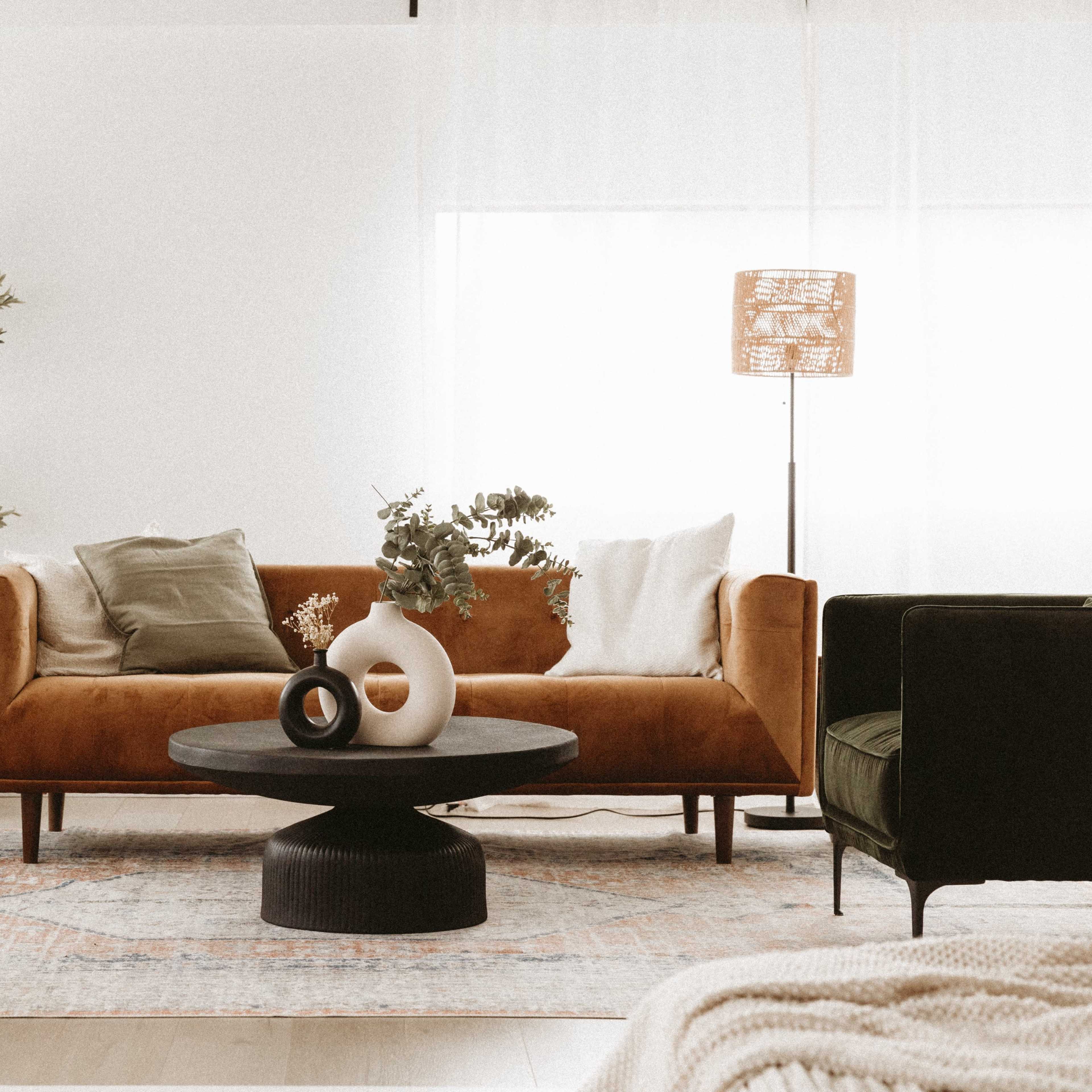 A modern living room with a brown velvet sofa, a round black coffee table, a green armchair, and decorative plants.