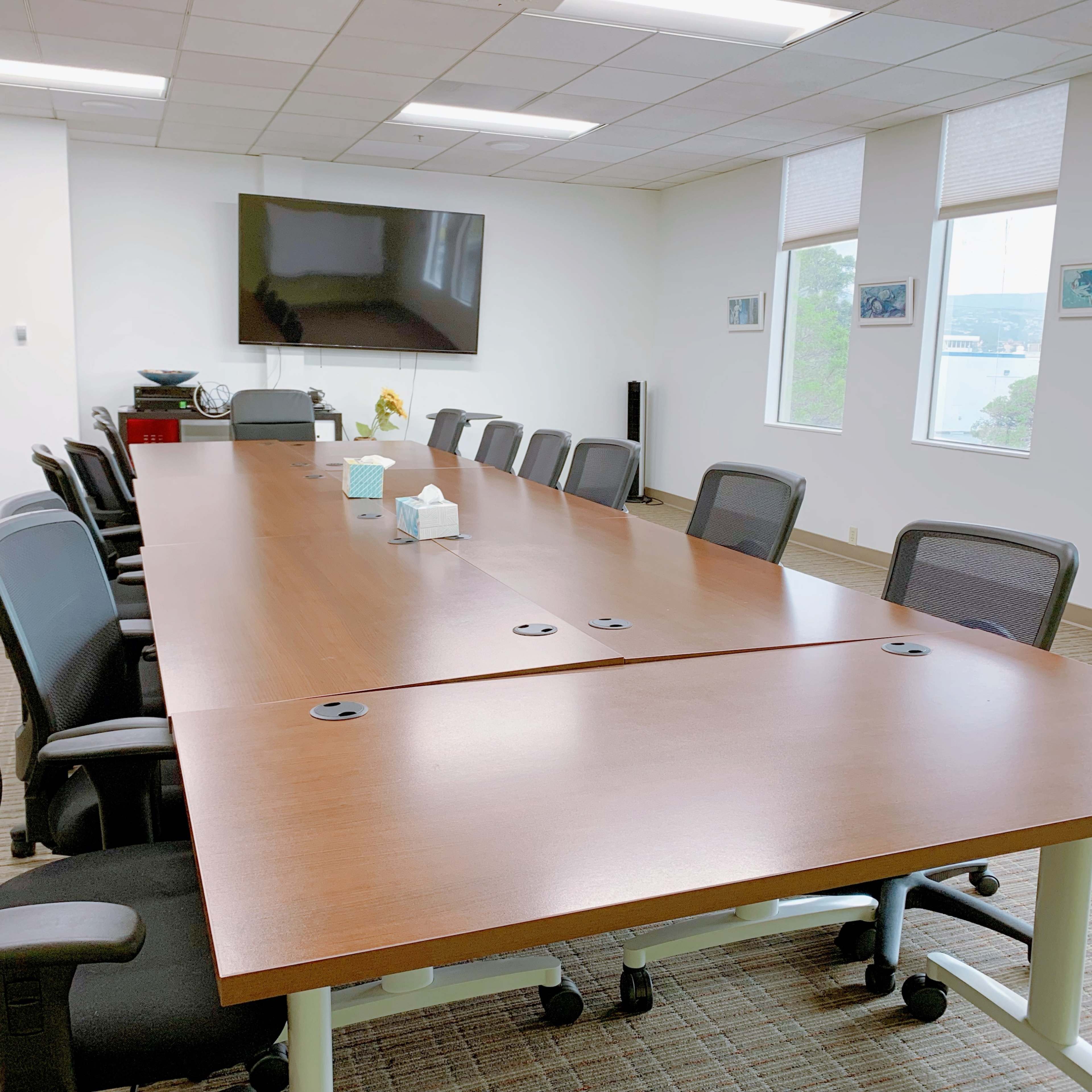 A long conference table surrounded by black office chairs in a brightly lit meeting room with a large screen on the wall.