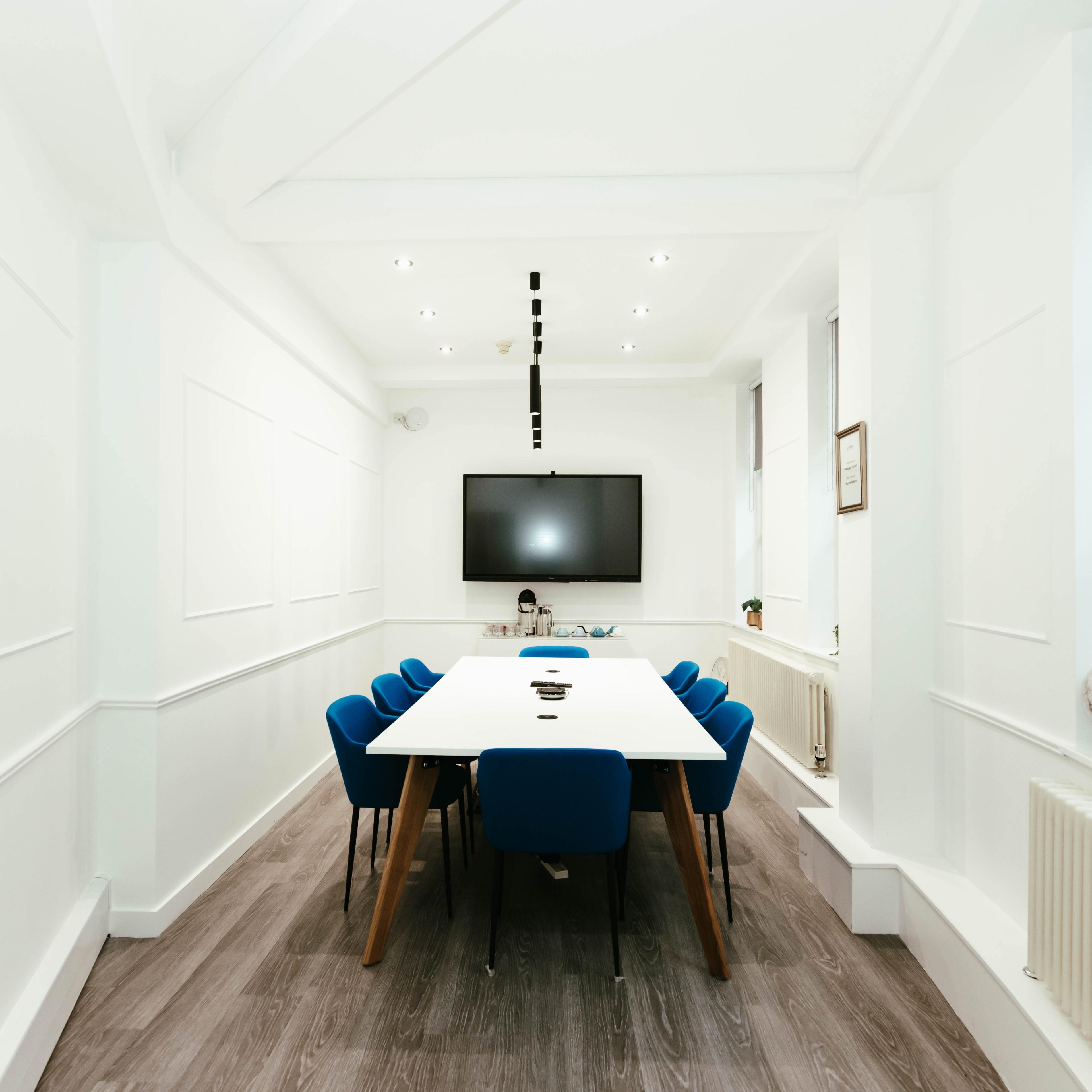 A modern conference room features a long white table surrounded by blue chairs, with a large flat-screen TV mounted on the wall.