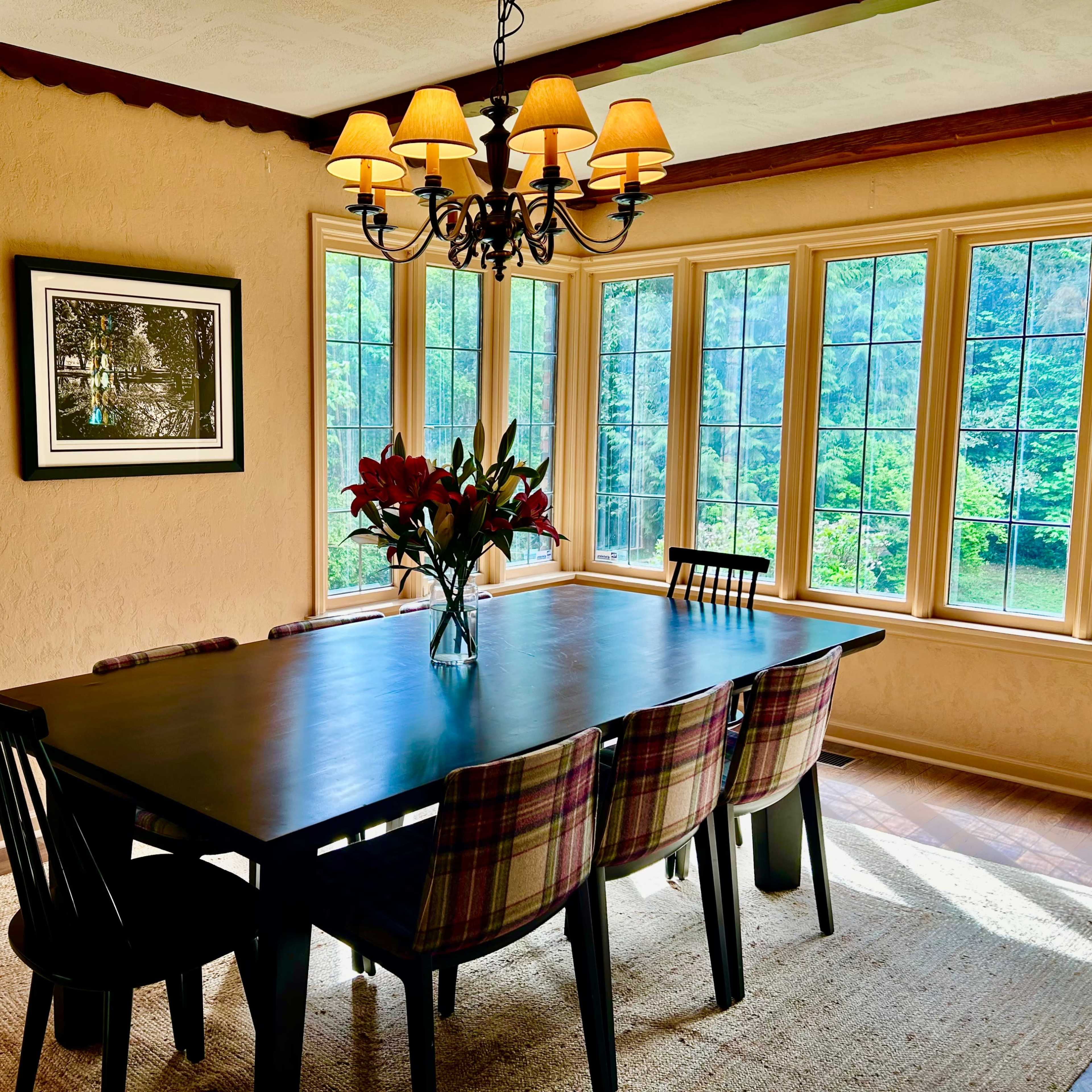 A dining room features a long black table surrounded by plaid chairs, illuminated by a chandelier, with large windows offering a view of greenery outside.