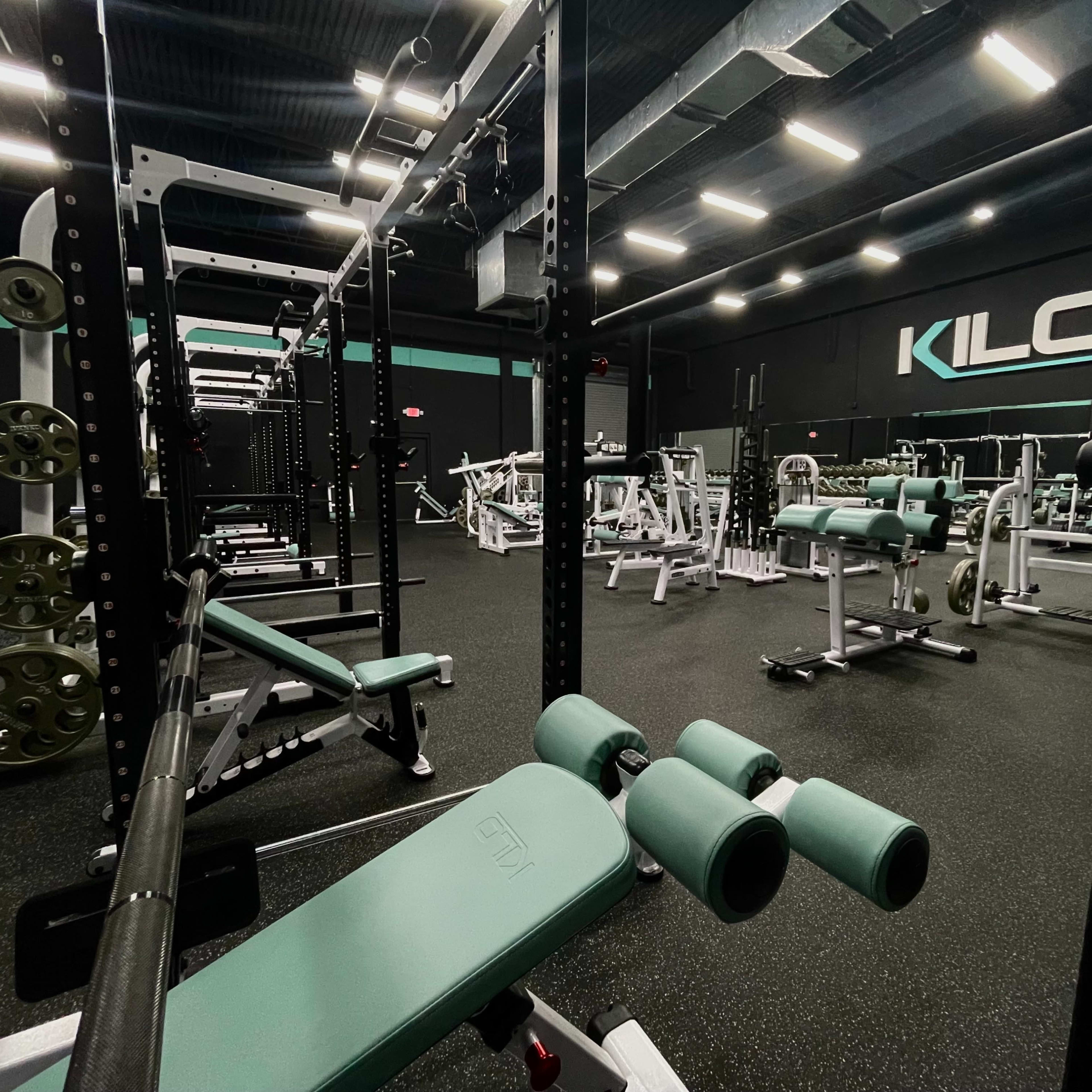 A well-equipped gym features various weightlifting machines and benches under bright overhead lighting.