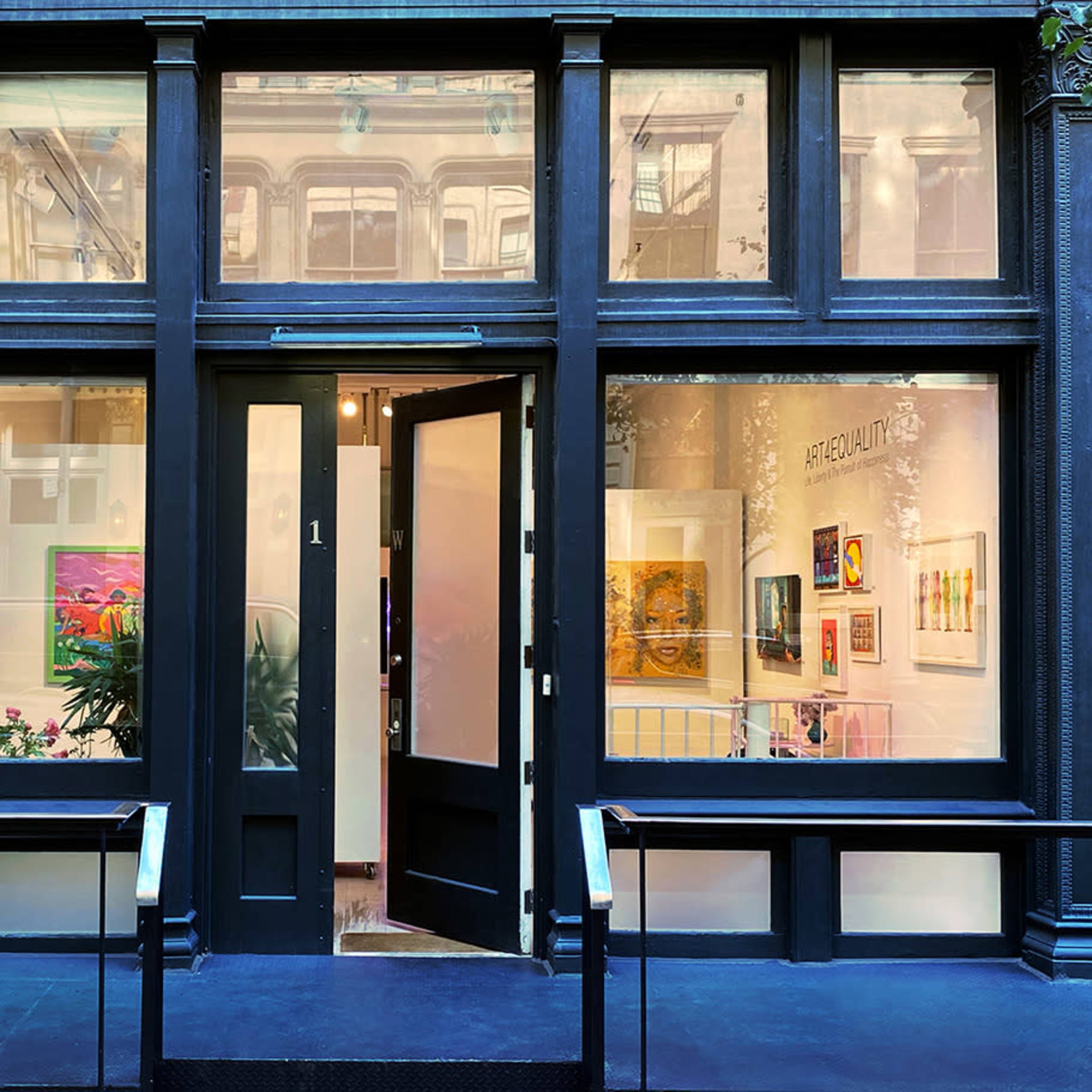 The entrance of an art gallery featuring large windows showcasing colorful artwork and lush plants.