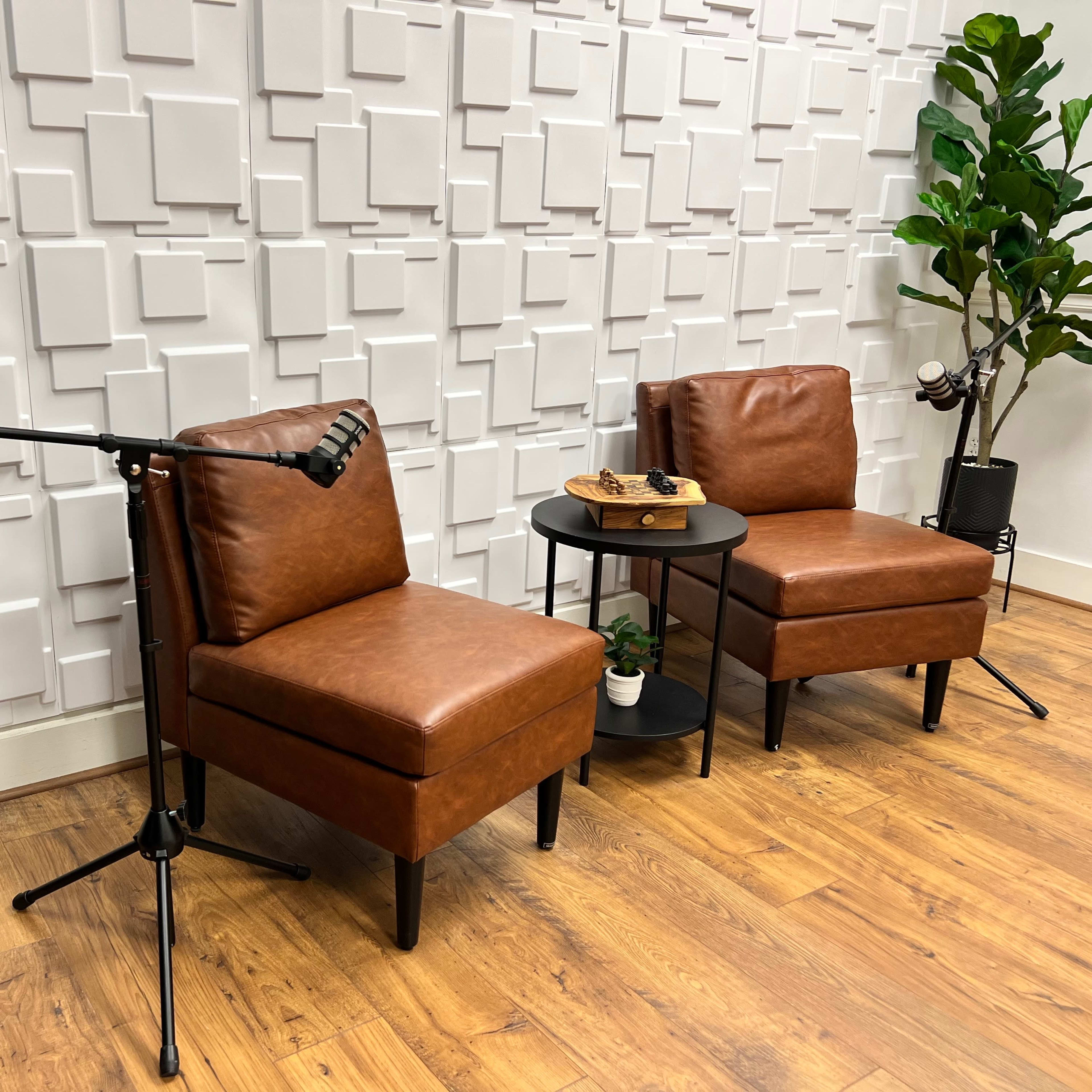 Two brown leather chairs positioned next to a small black table with a plate of snacks, surrounded by decorative plants against a textured white wall.
