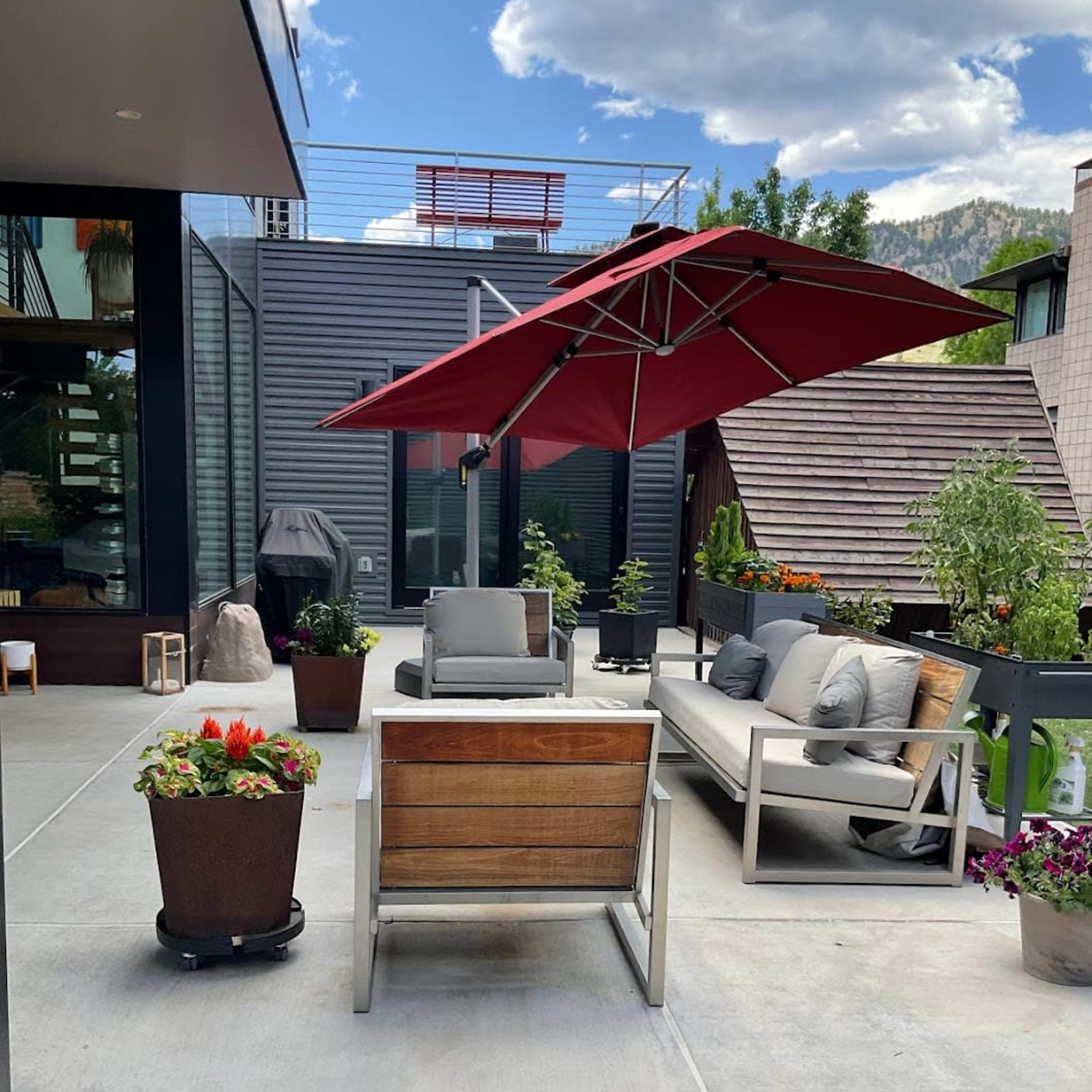 A modern outdoor patio with a red umbrella, seating areas, and potted plants against a backdrop of buildings and mountains.