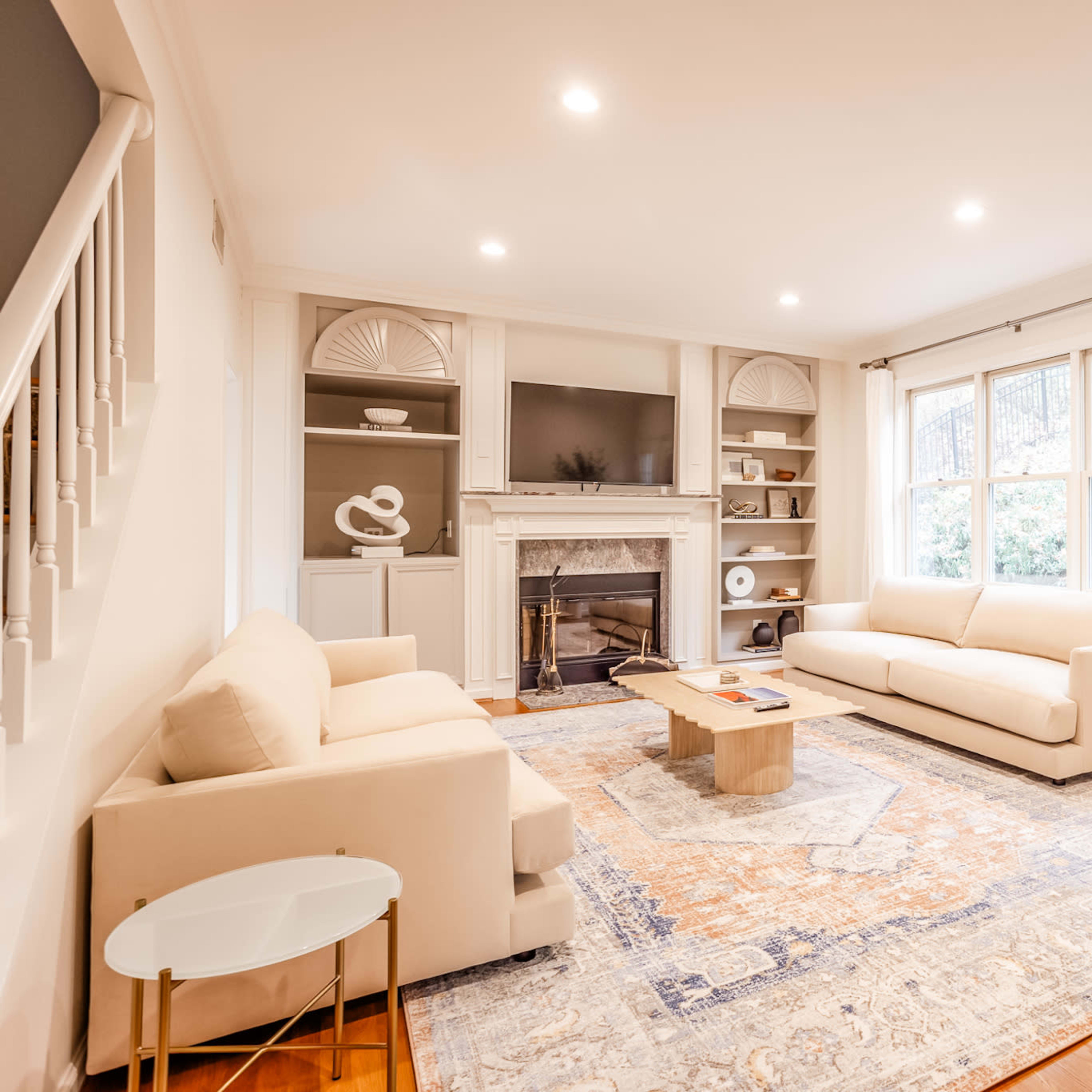 A bright living room features two beige sofas, a wooden coffee table, and a fireplace, with a large area rug and windows allowing natural light to enter.