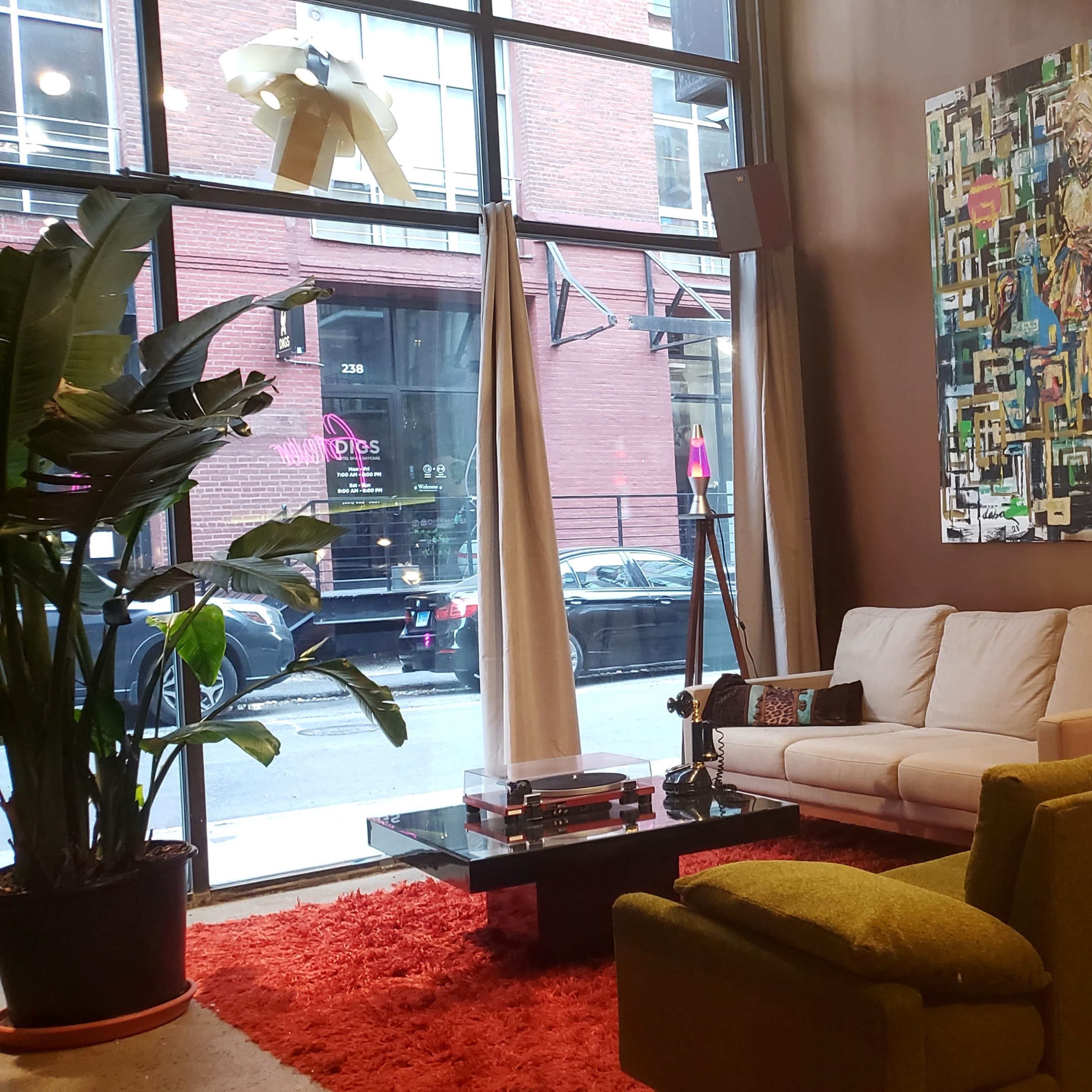 The interior of a modern lounge features a white couch and green armchair facing a coffee table, with large windows showcasing an urban street view and artwork on the walls.