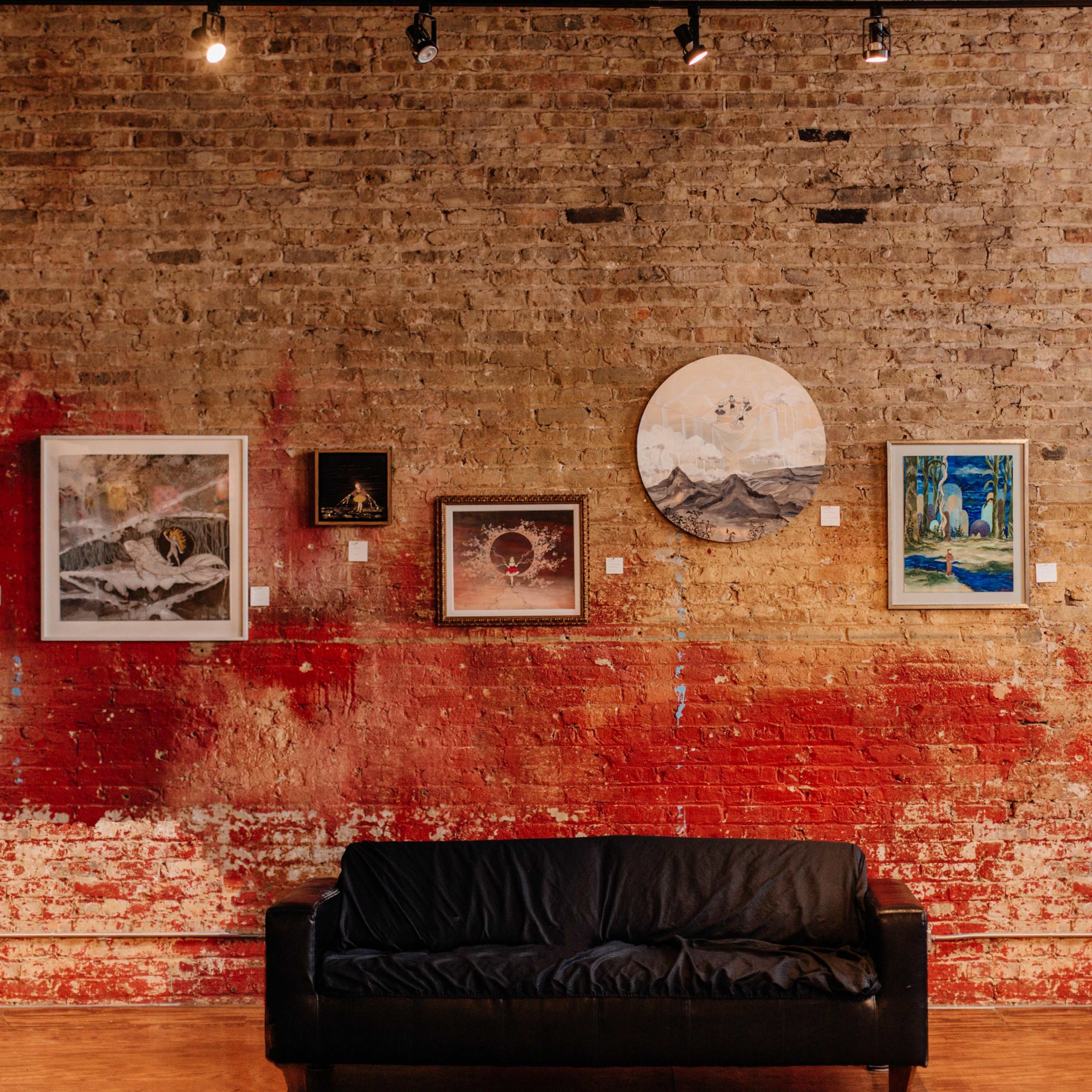 A gallery wall featuring various framed artworks displayed against a brick wall with a splash of red paint, alongside a black couch in the foreground.