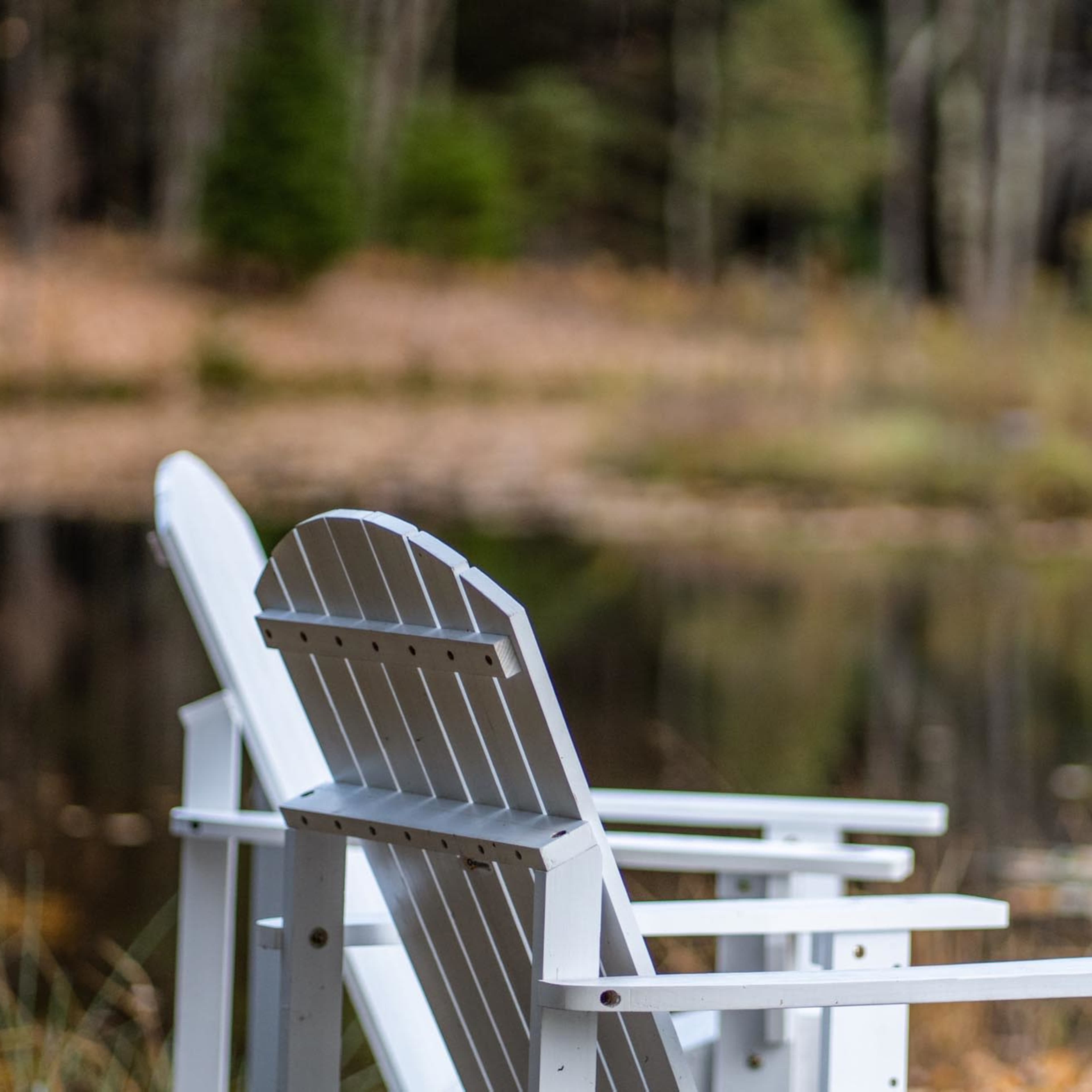 Two white wooden chairs positioned near a pond surrounded by trees.