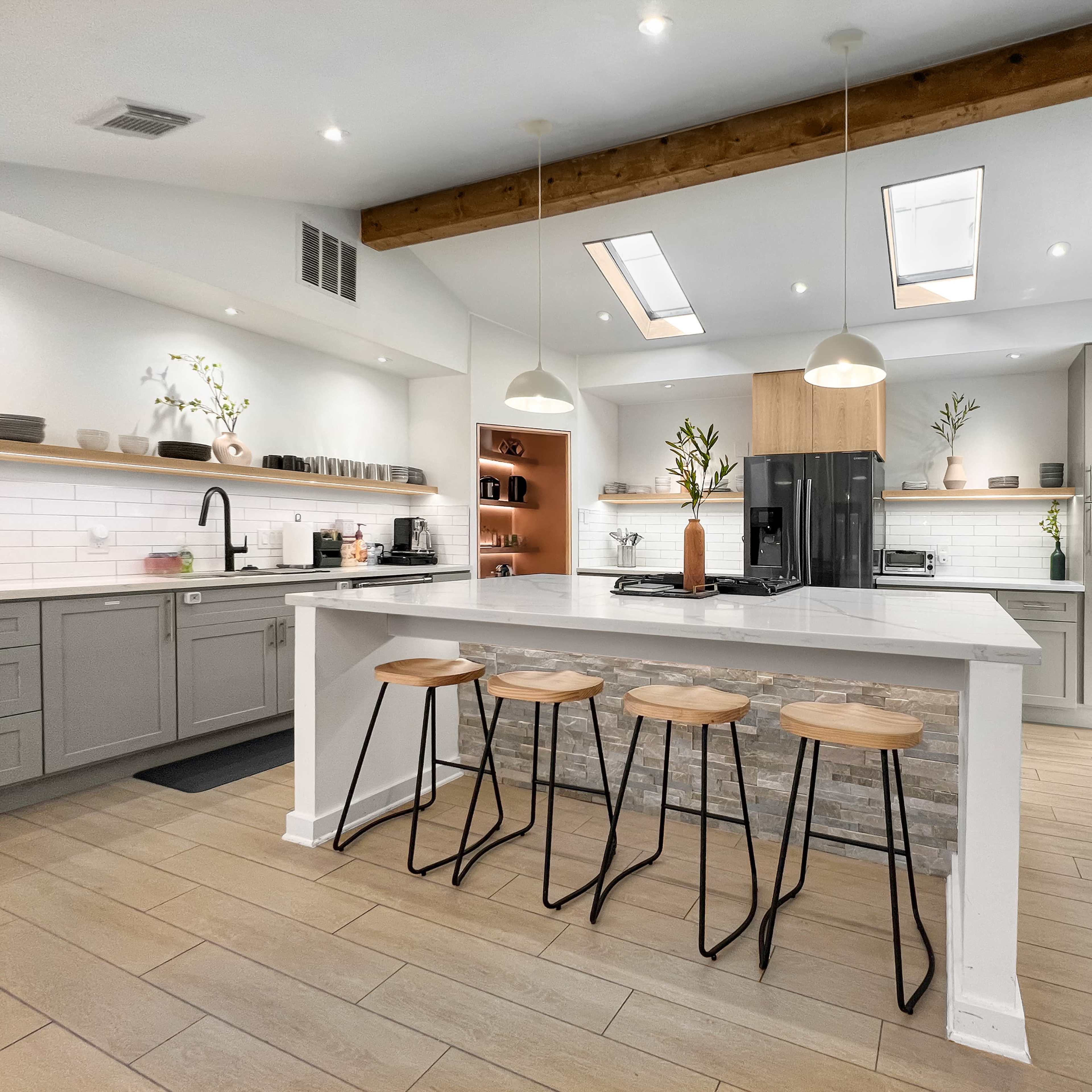 A modern kitchen features a central island with four wooden stools, sleek cabinetry, and natural light streaming through skylights.