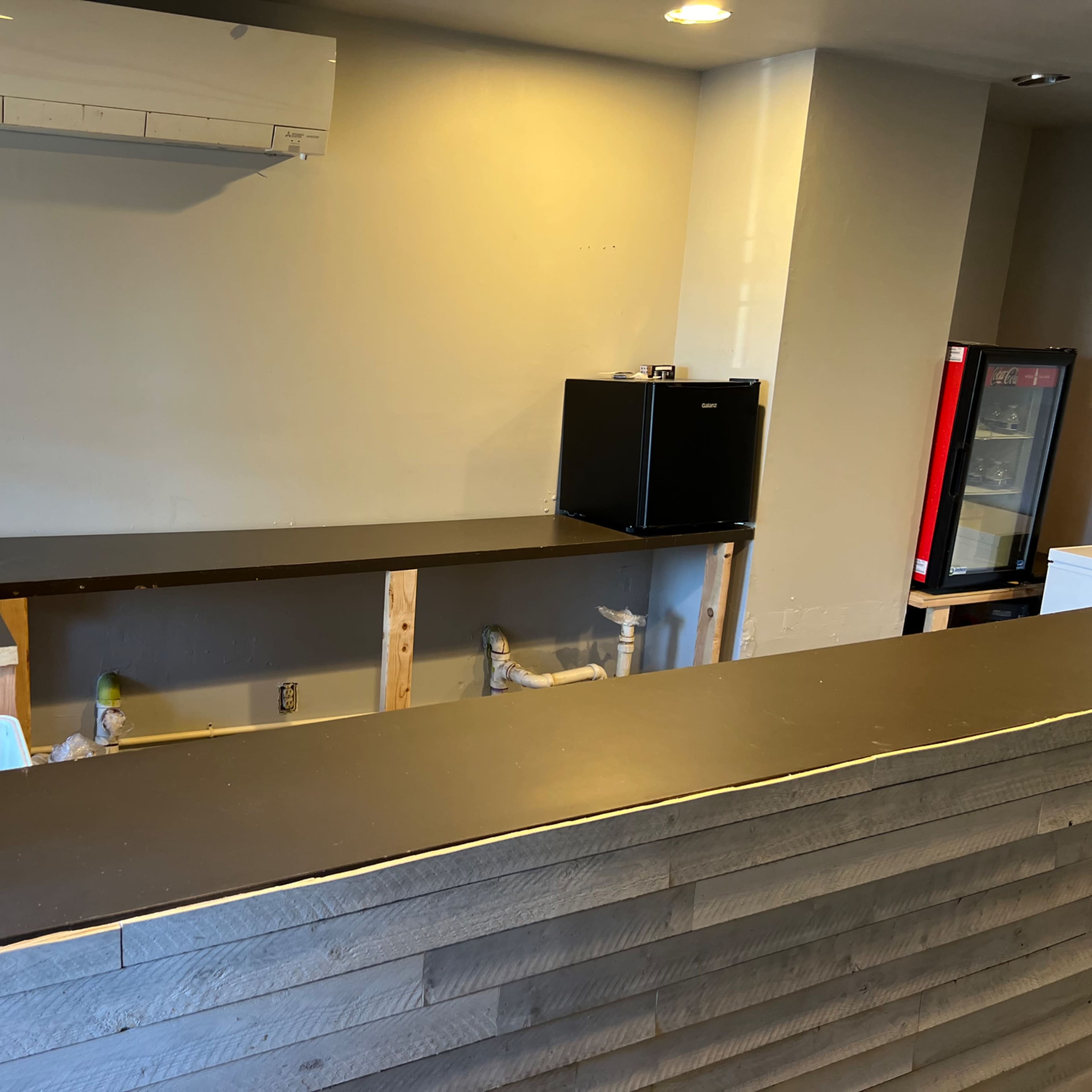 A partially finished counter area with a sink, refrigerator, and plumbing setup in a room featuring plain walls and ceiling.