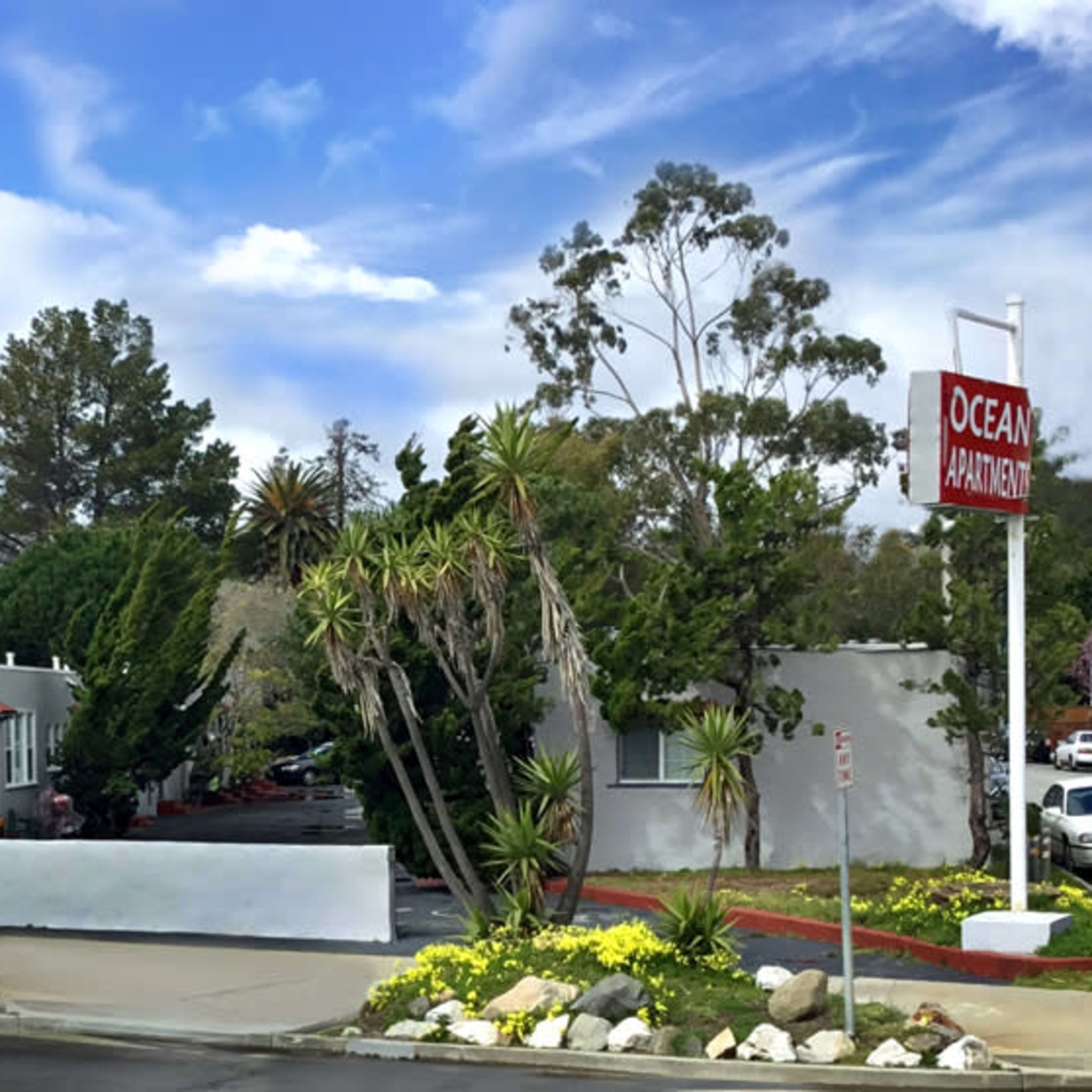 A row of apartment buildings with landscaped greenery and palm trees under a blue sky.