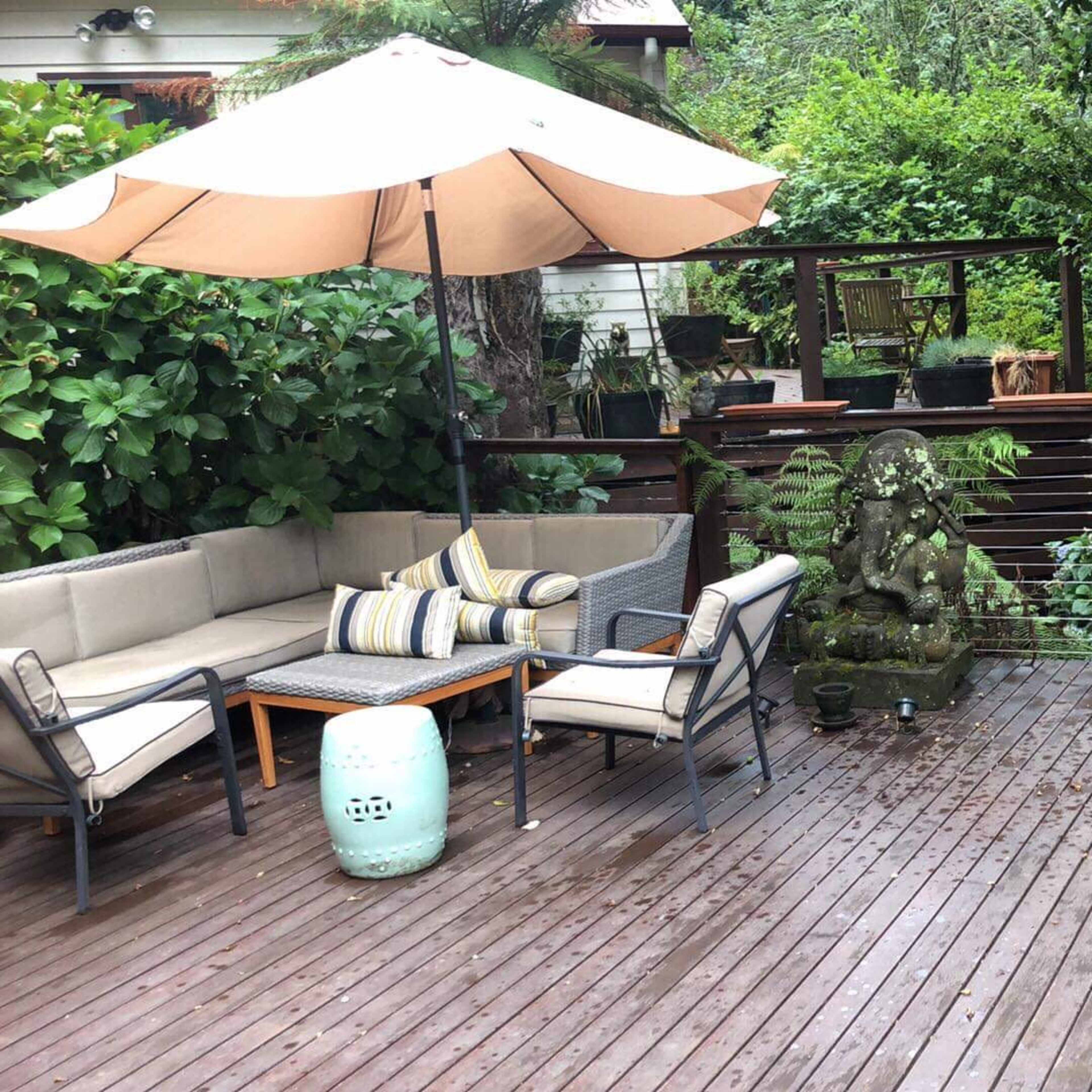 A wooden deck area with a seating arrangement under an umbrella, a small stone statue, and a dining table surrounded by lush greenery.