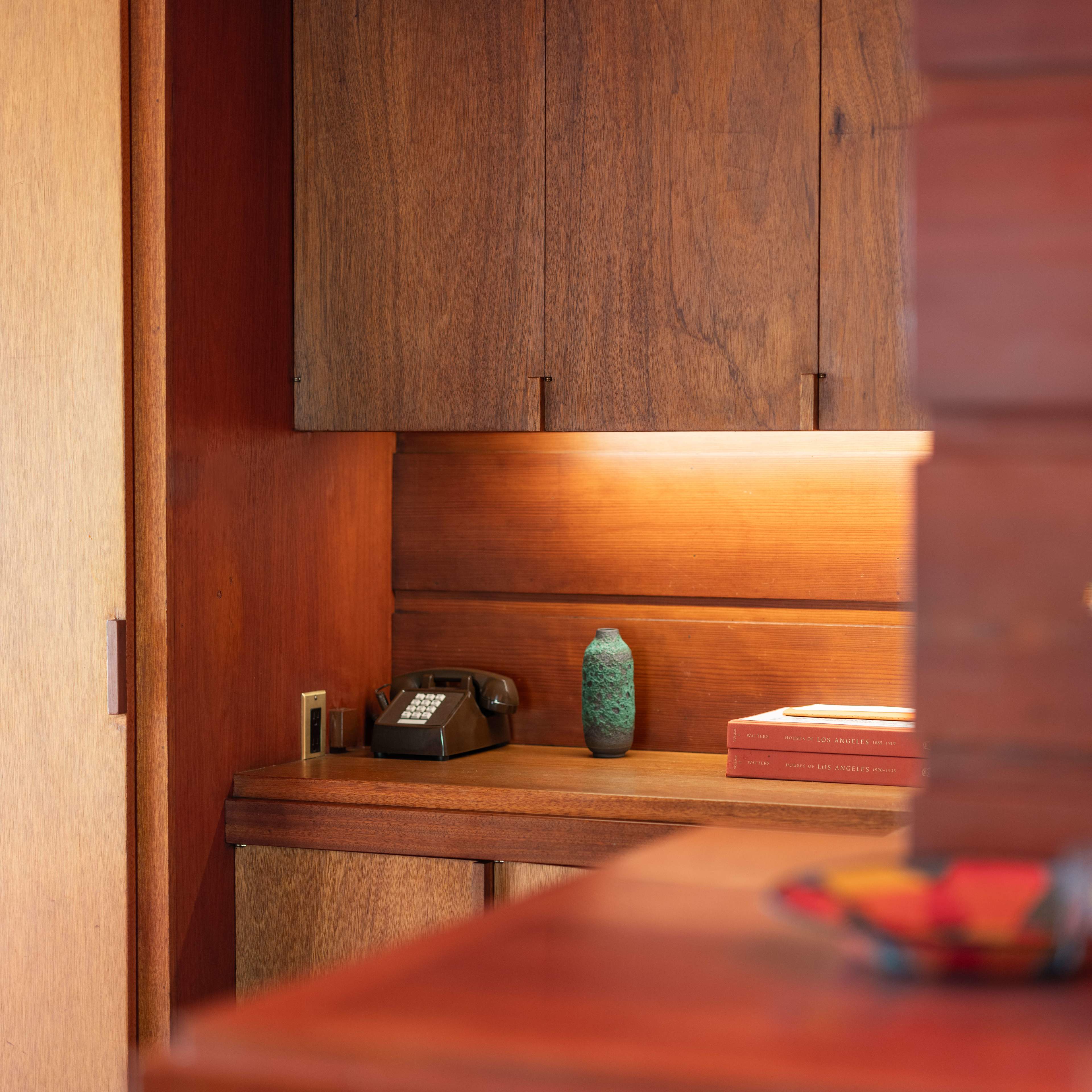 A wooden interior with a small desk area featuring a vintage telephone, a decorative object, and a stack of books illuminated by soft lighting.
