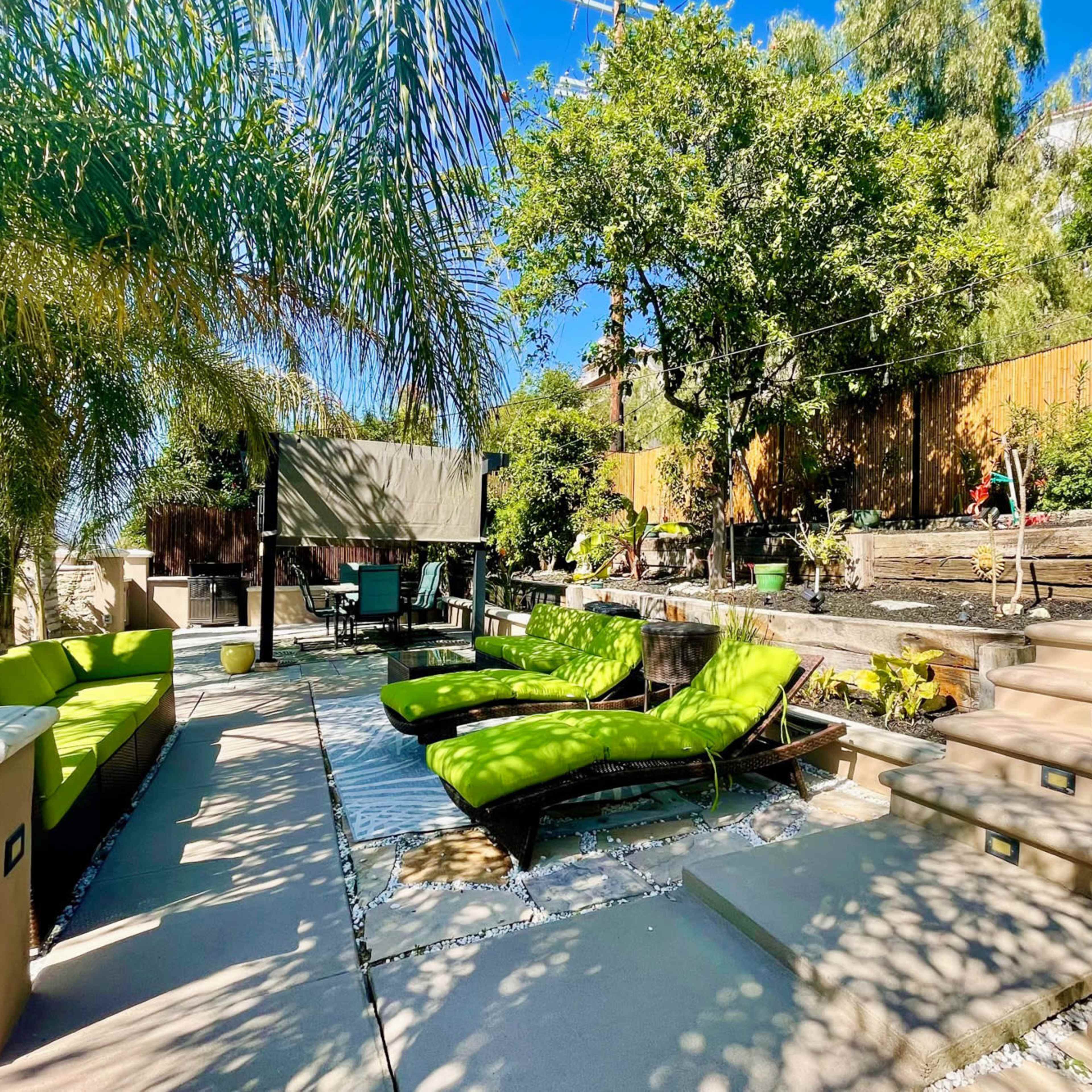 A landscaped outdoor patio area with green lounge chairs, a dining set, and tropical plants under a clear blue sky.