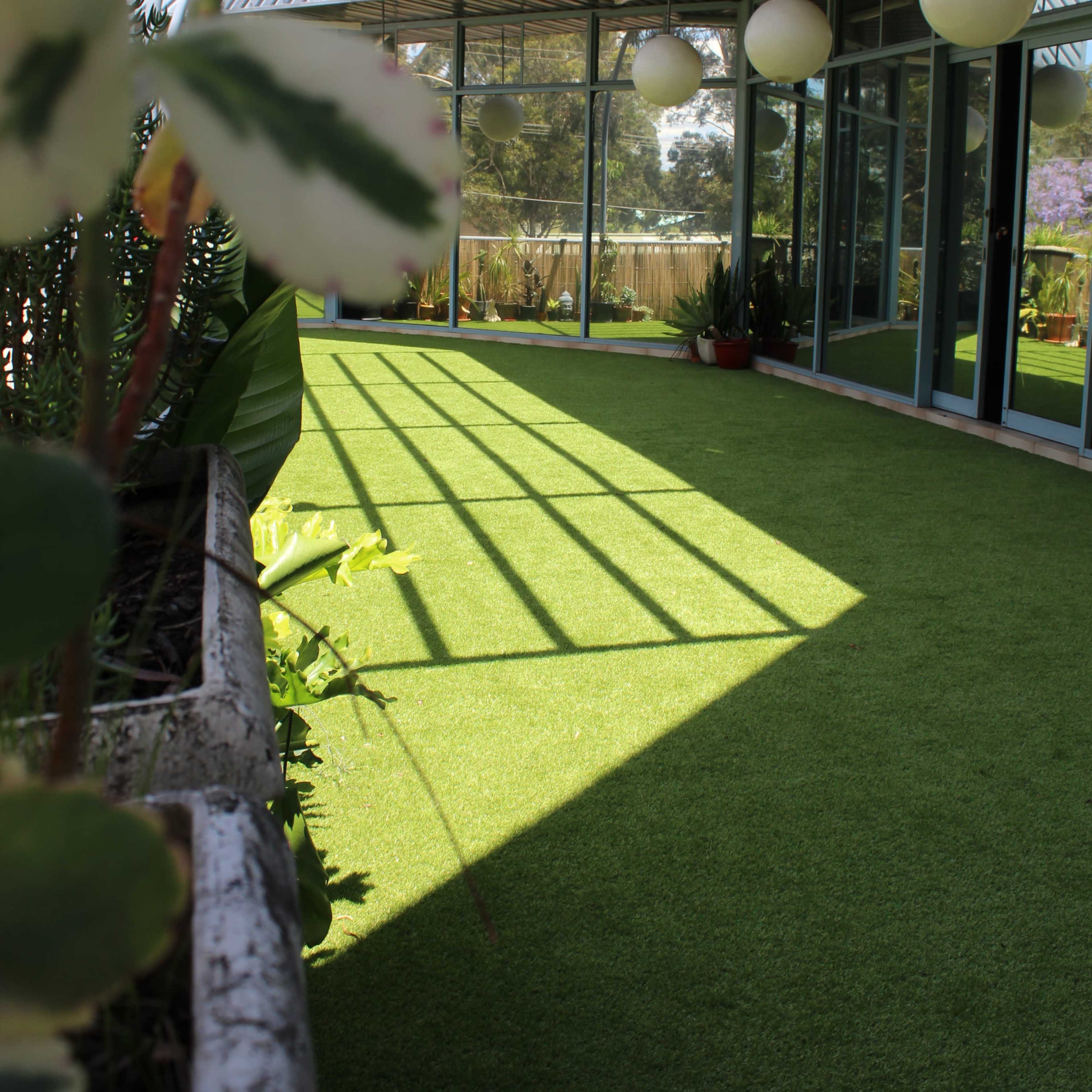 A glass-enclosed space with artificial turf flooring, featuring potted plants and suspended light fixtures.