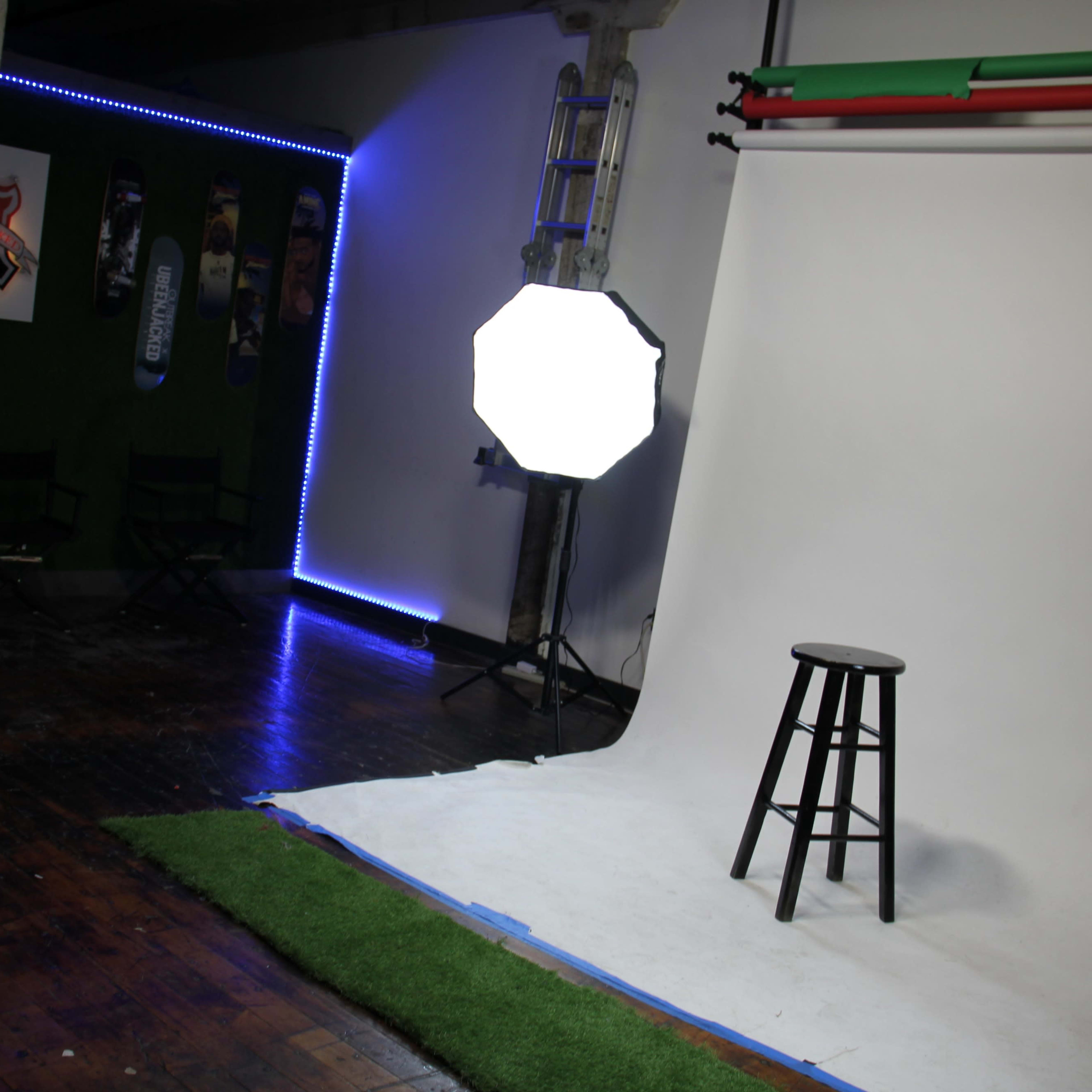 A photography studio with a white backdrop, a stool, and lighting equipment, complemented by green turf on the ground and neon blue accents along the walls.