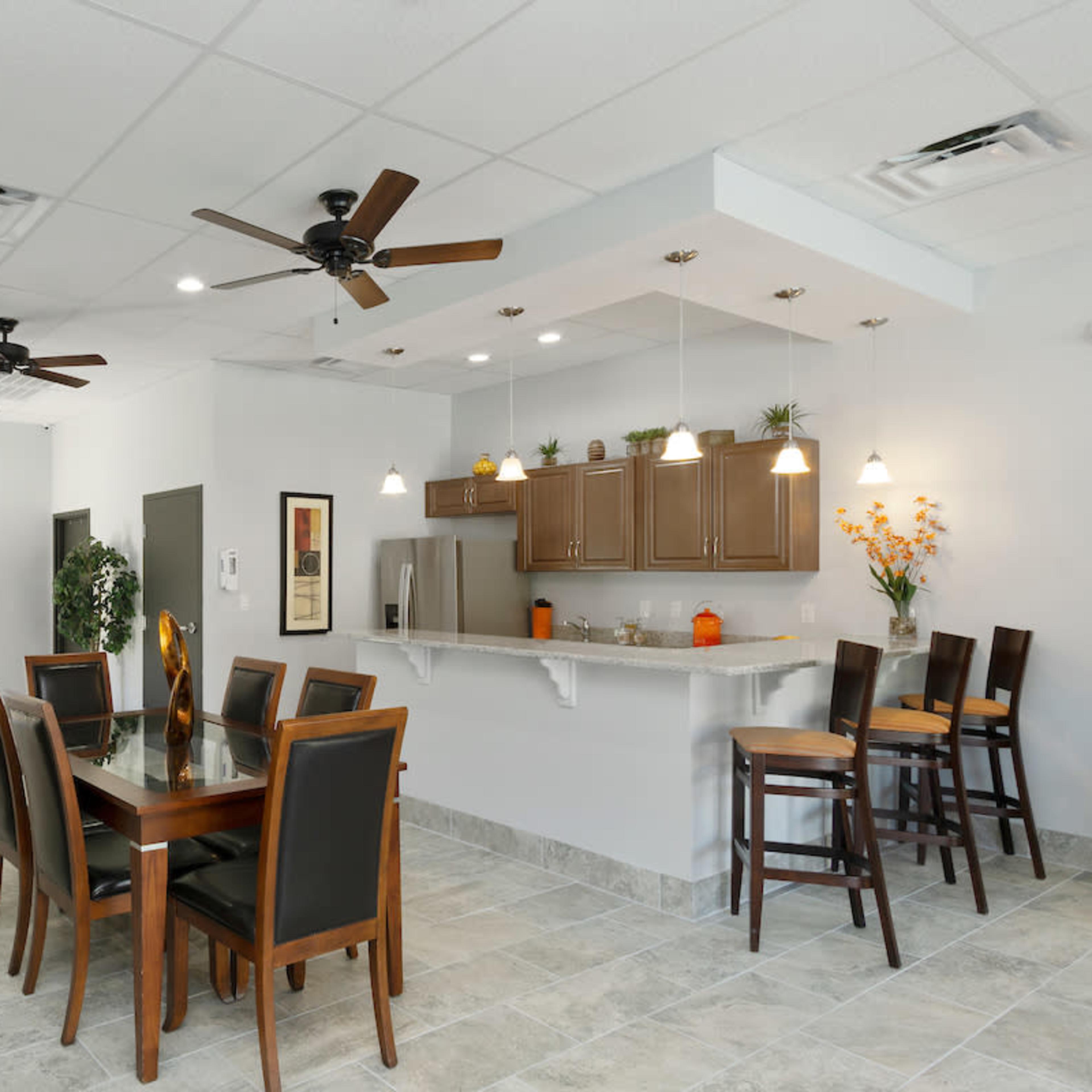A modern dining area with a wooden table and chairs, a breakfast bar, and kitchen cabinetry, all under ceiling fans and bright lighting.