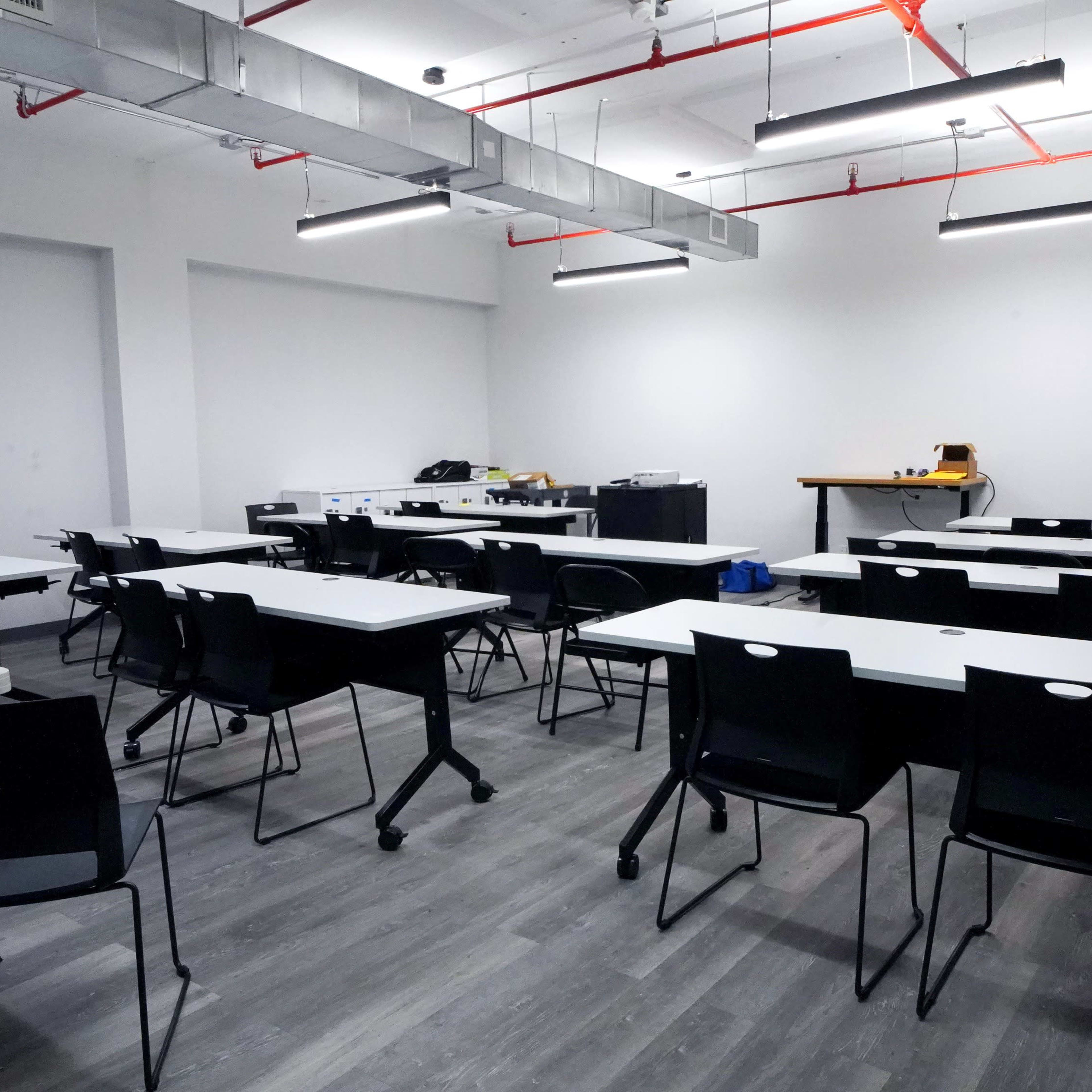 A nearly empty classroom with rows of black chairs and white tables, illuminated by overhead lights.