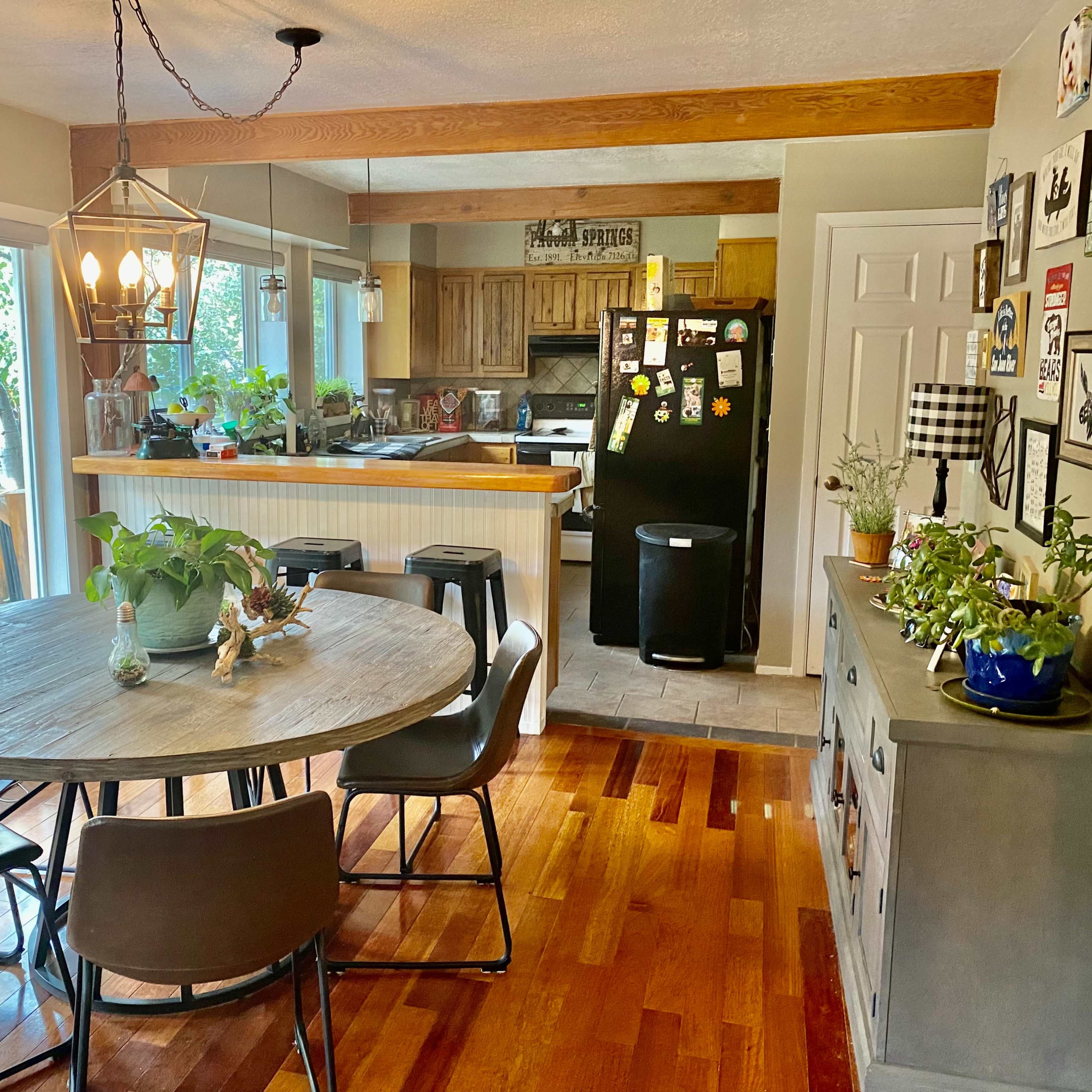 A round dining table surrounded by chairs is positioned in a bright kitchen area with wooden floors and a view of the outdoors through large windows.