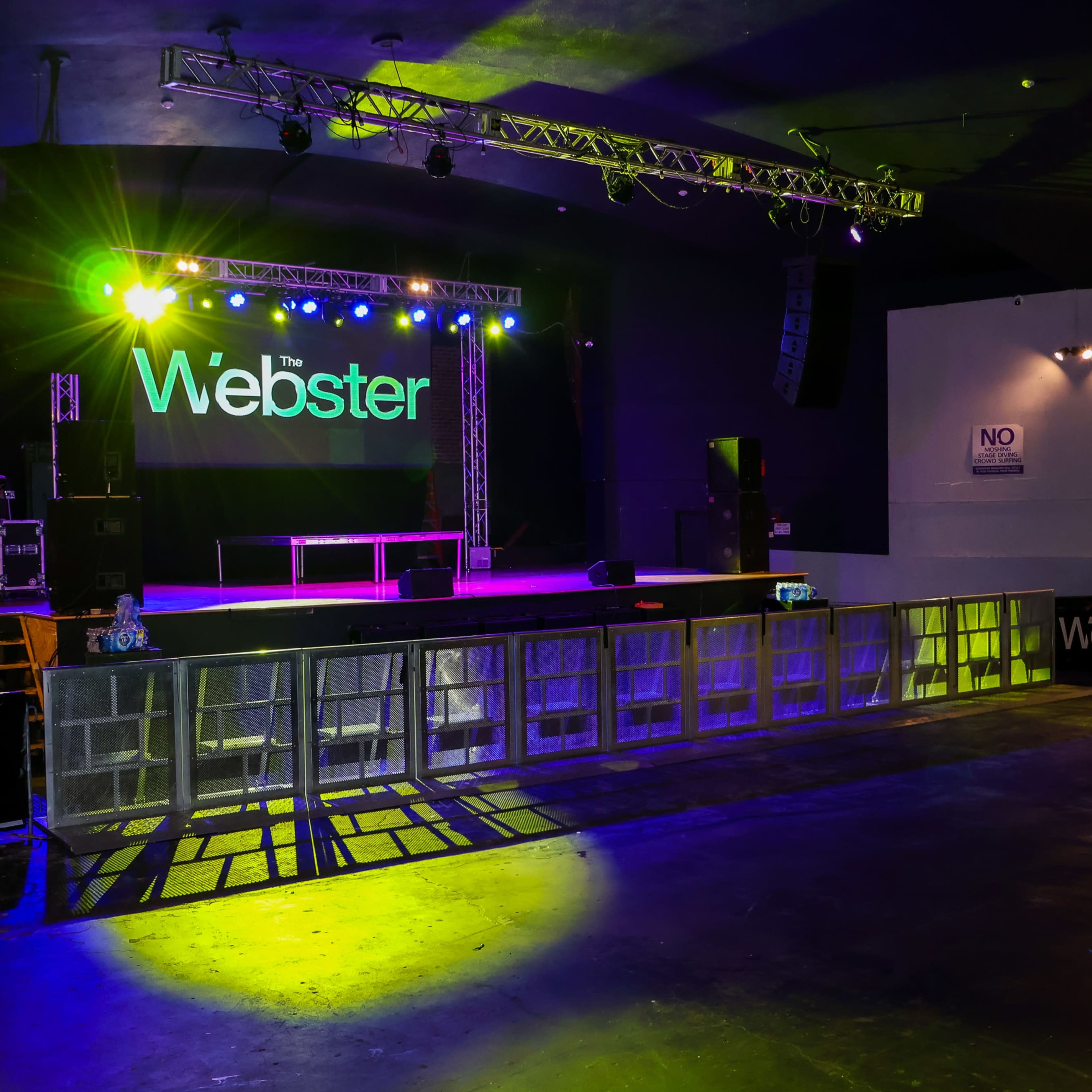 A small concert venue with a stage illuminated by colorful lights, featuring barriers in front of the stage and signage displaying "the Webster."