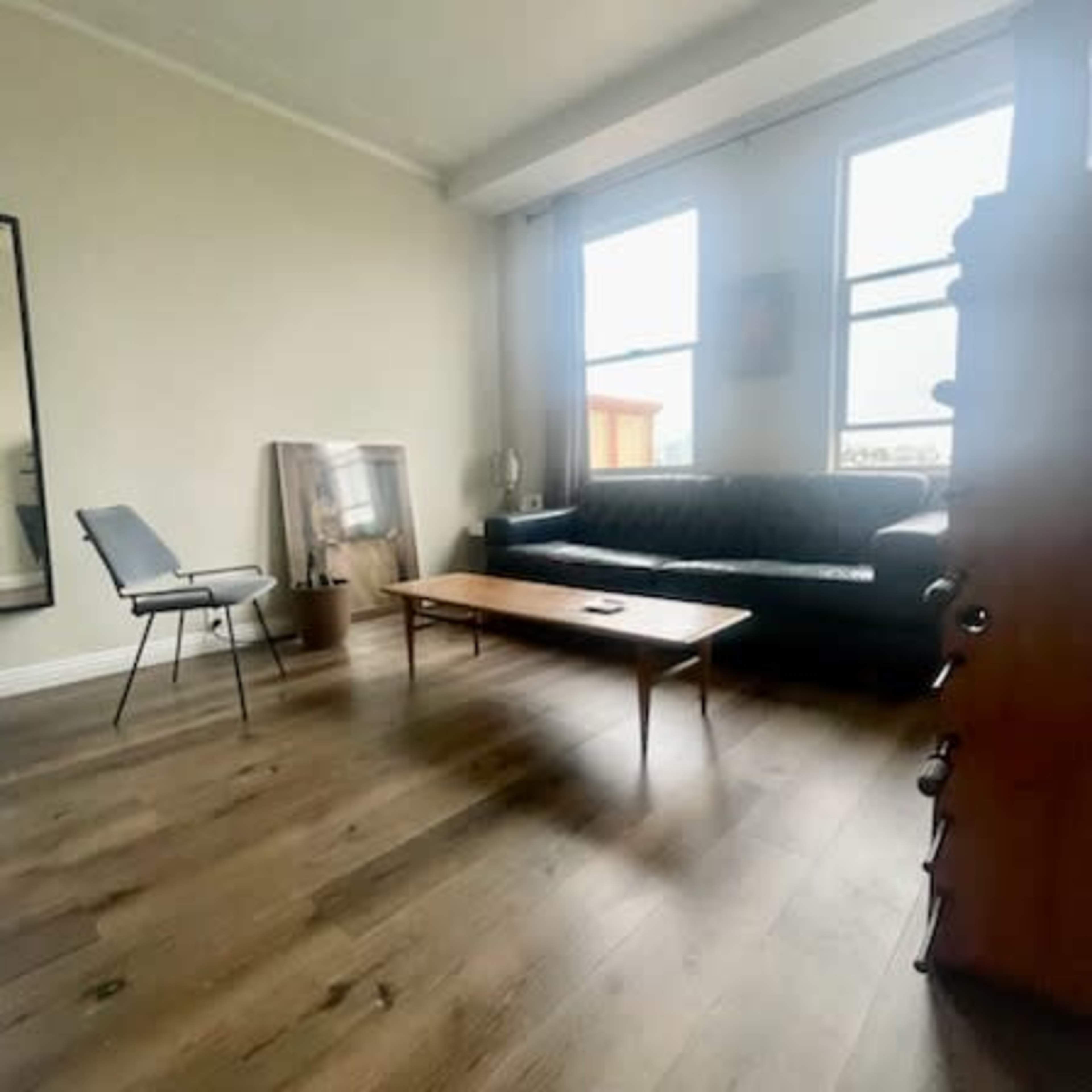 The room features a black sofa facing a wooden coffee table, with a chair nearby and large windows letting in natural light.