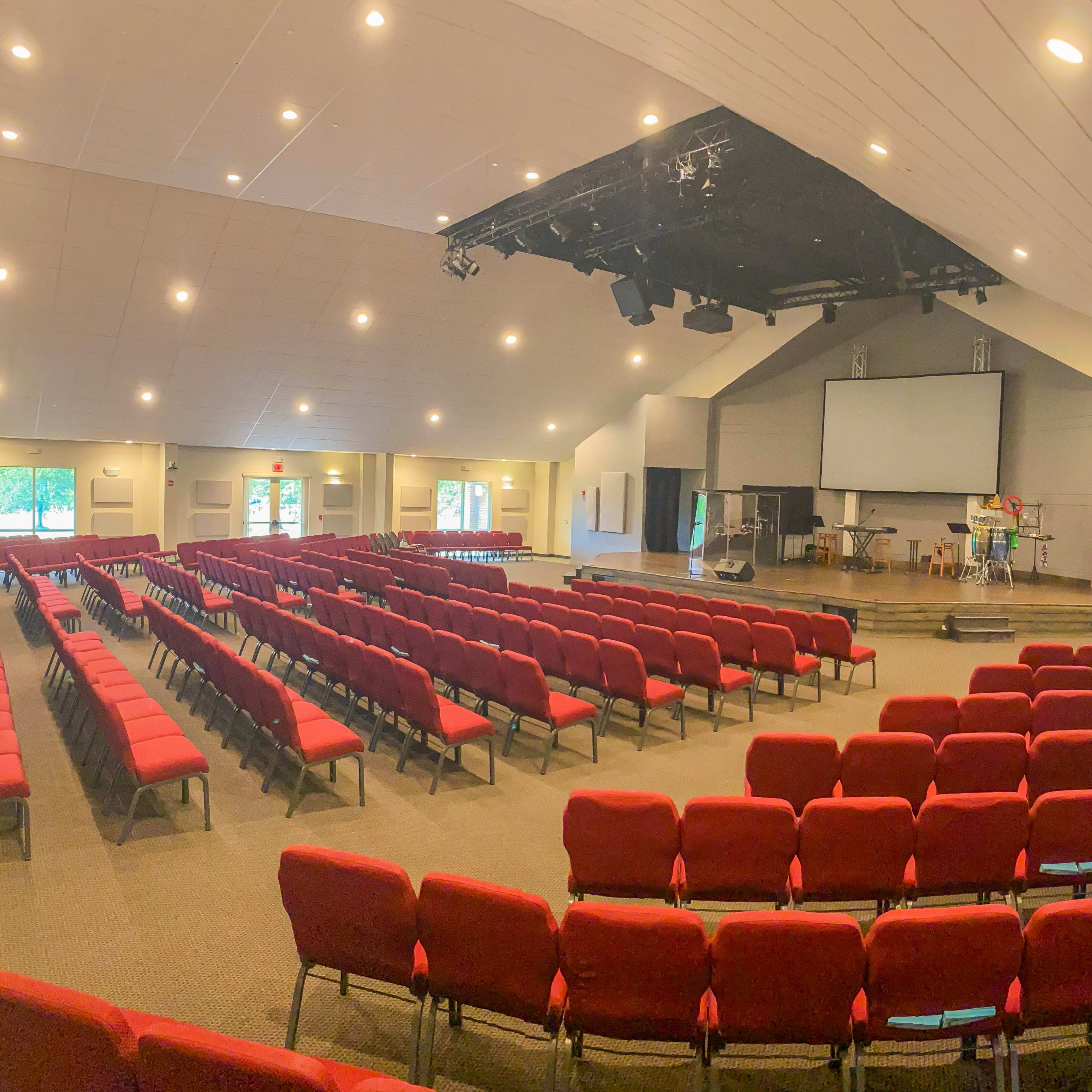 A large, empty auditorium with rows of red chairs facing a stage equipped with sound equipment and a projector screen.