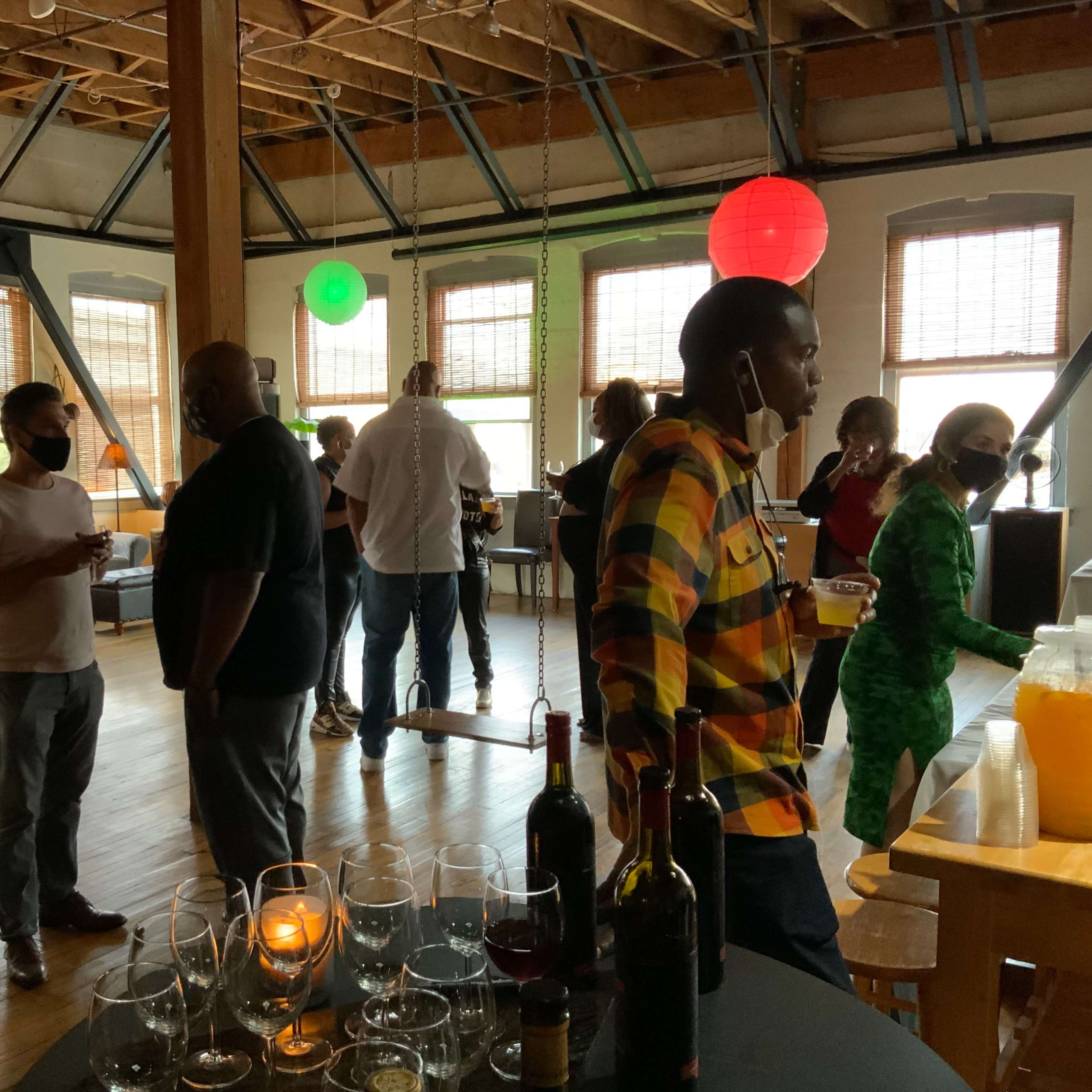 People are socializing at a gathering in a spacious room with colorful lanterns and a table of drinks.