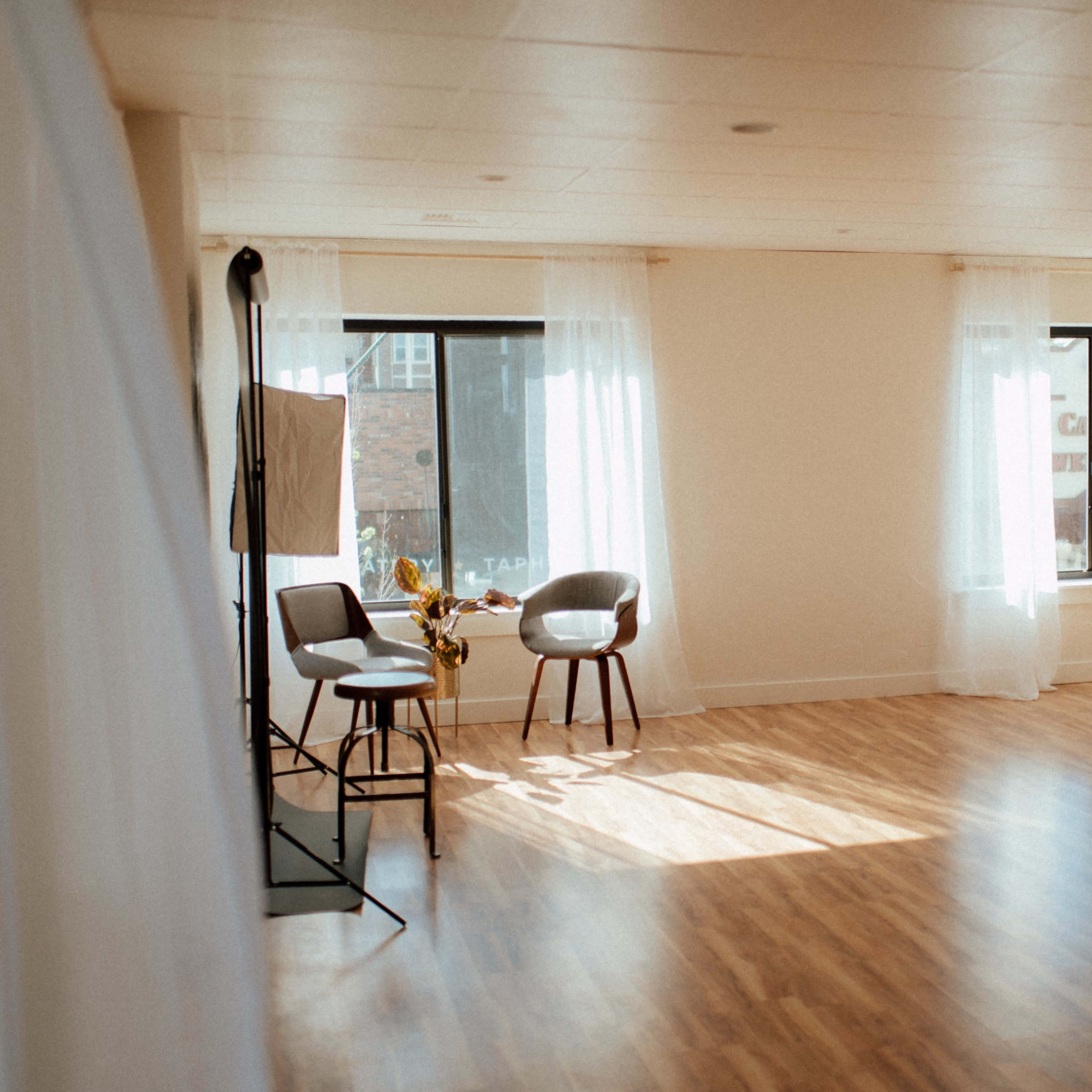 A bright and open studio space features two chairs, large windows with sheer curtains, and a potted plant.