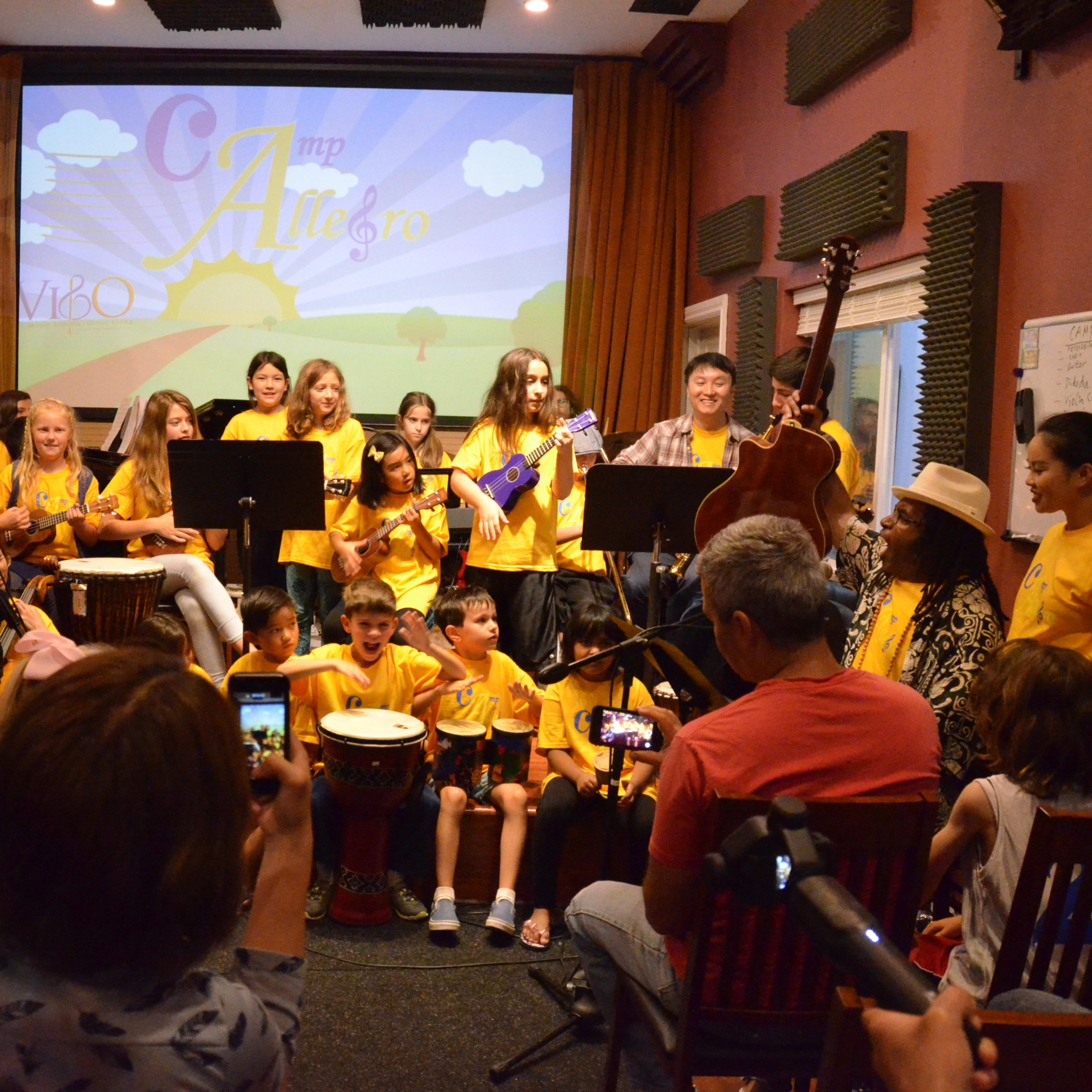 A large group of children in yellow shirts plays various musical instruments in a performance setting, while an audience records the event on their devices.