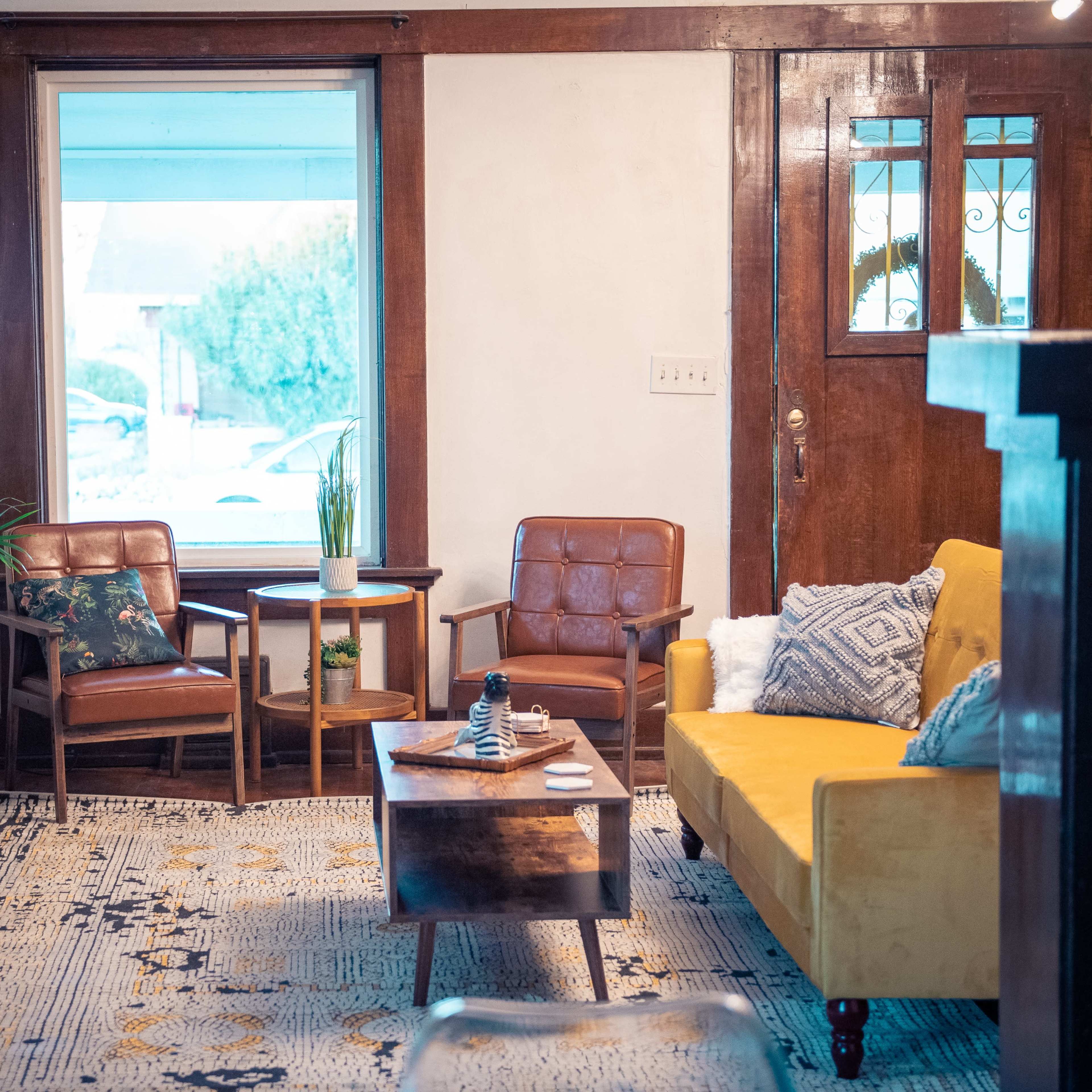 The room features a mix of brown leather chairs, a yellow sofa, a wooden coffee table, and a large window with a view of the outside.