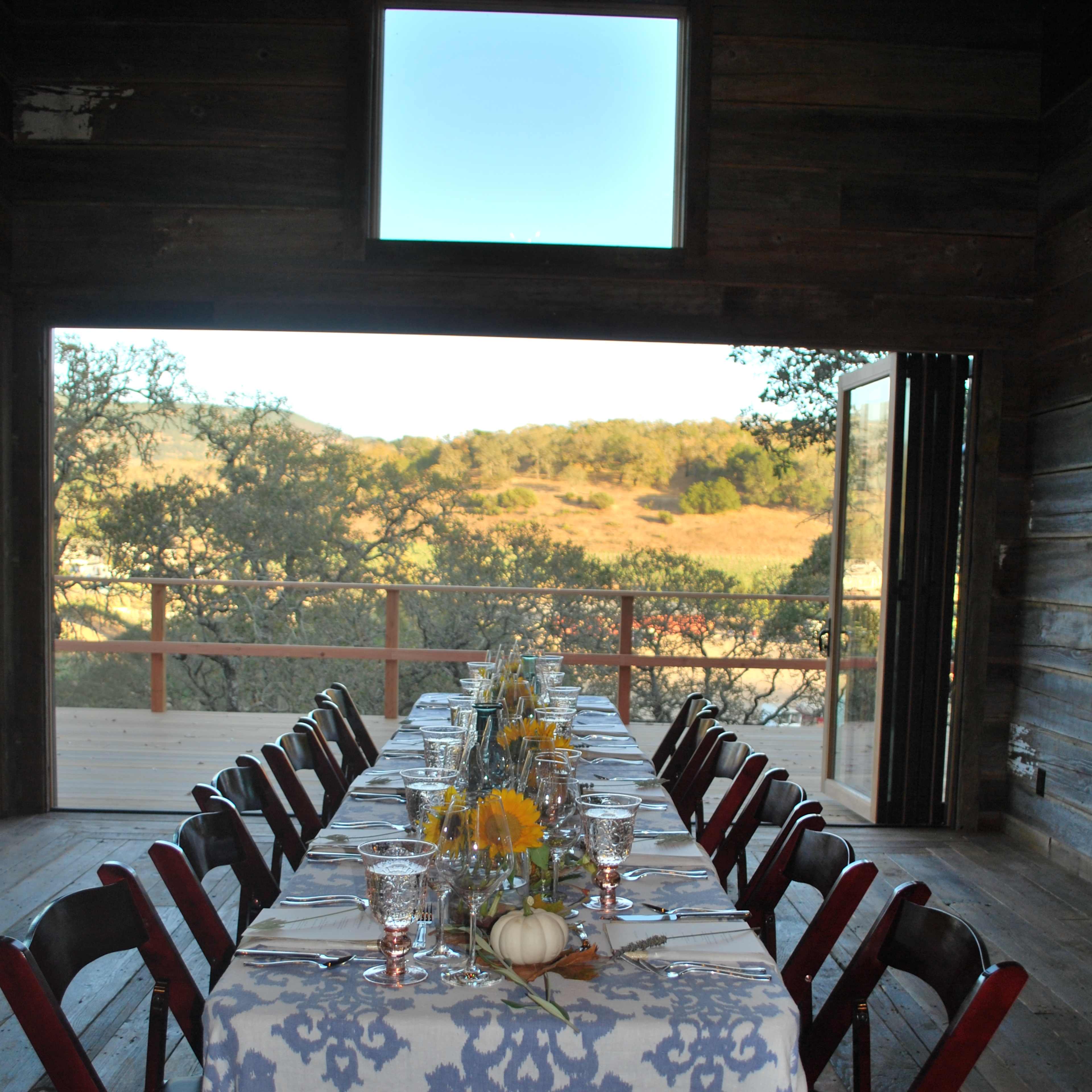 A long dining table is set with a floral centerpiece and is positioned in front of an open doorway that leads to a view of a hillside.