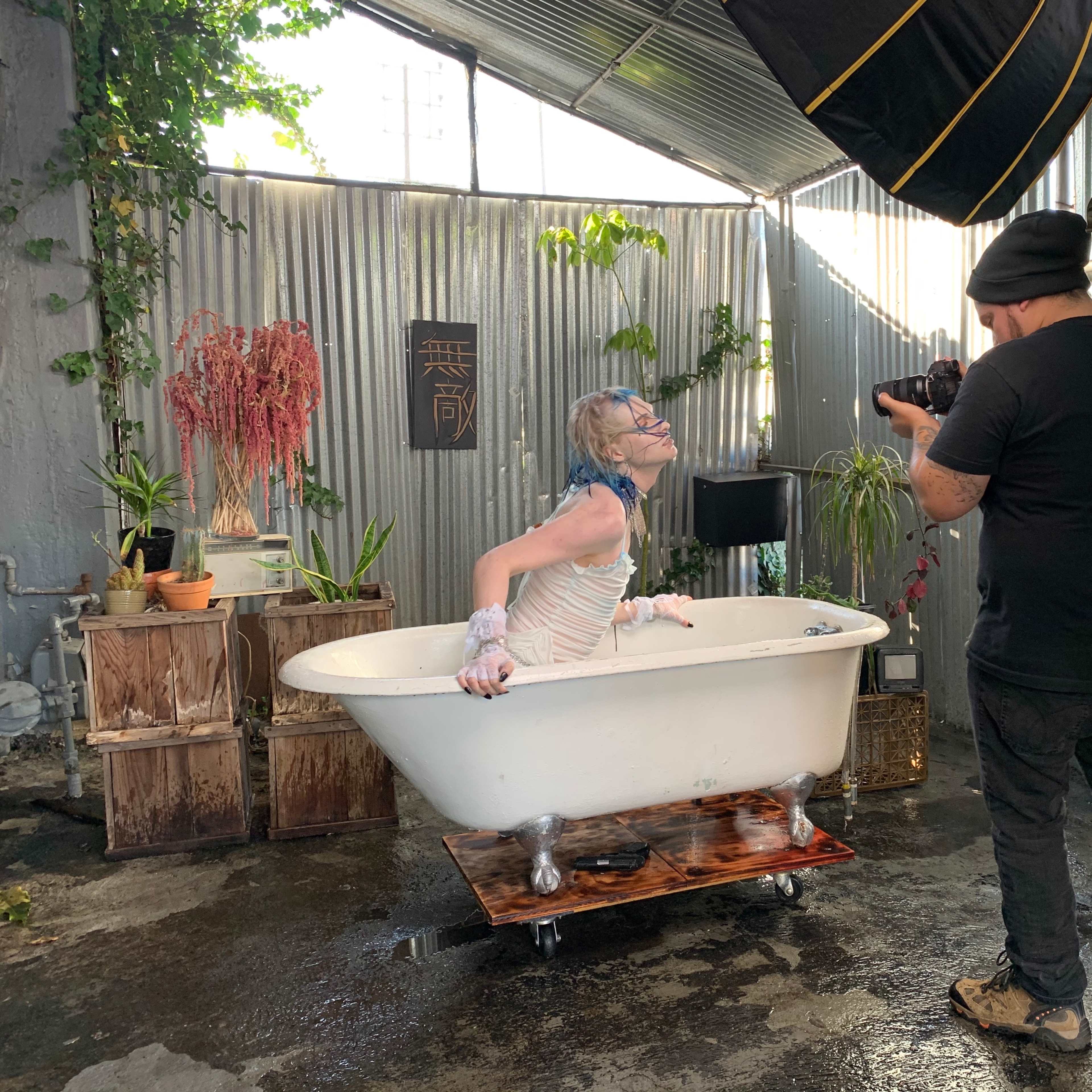 A person is emerging from a bathtub while two photographers capture the scene in an industrial-style setting with greenery and metal walls.