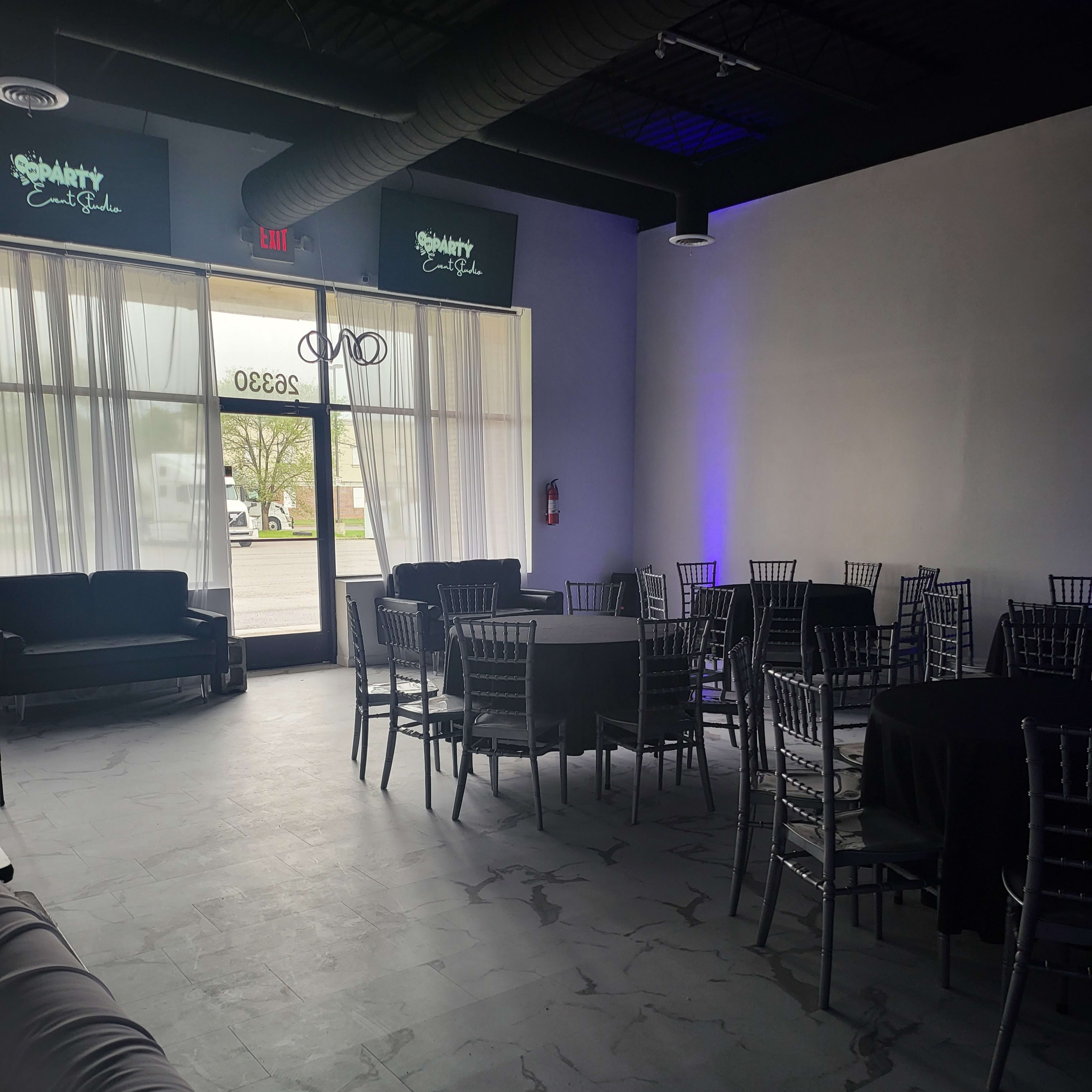 A spacious event venue with black tables and silver chiavari chairs, illuminated by natural light from large windows.