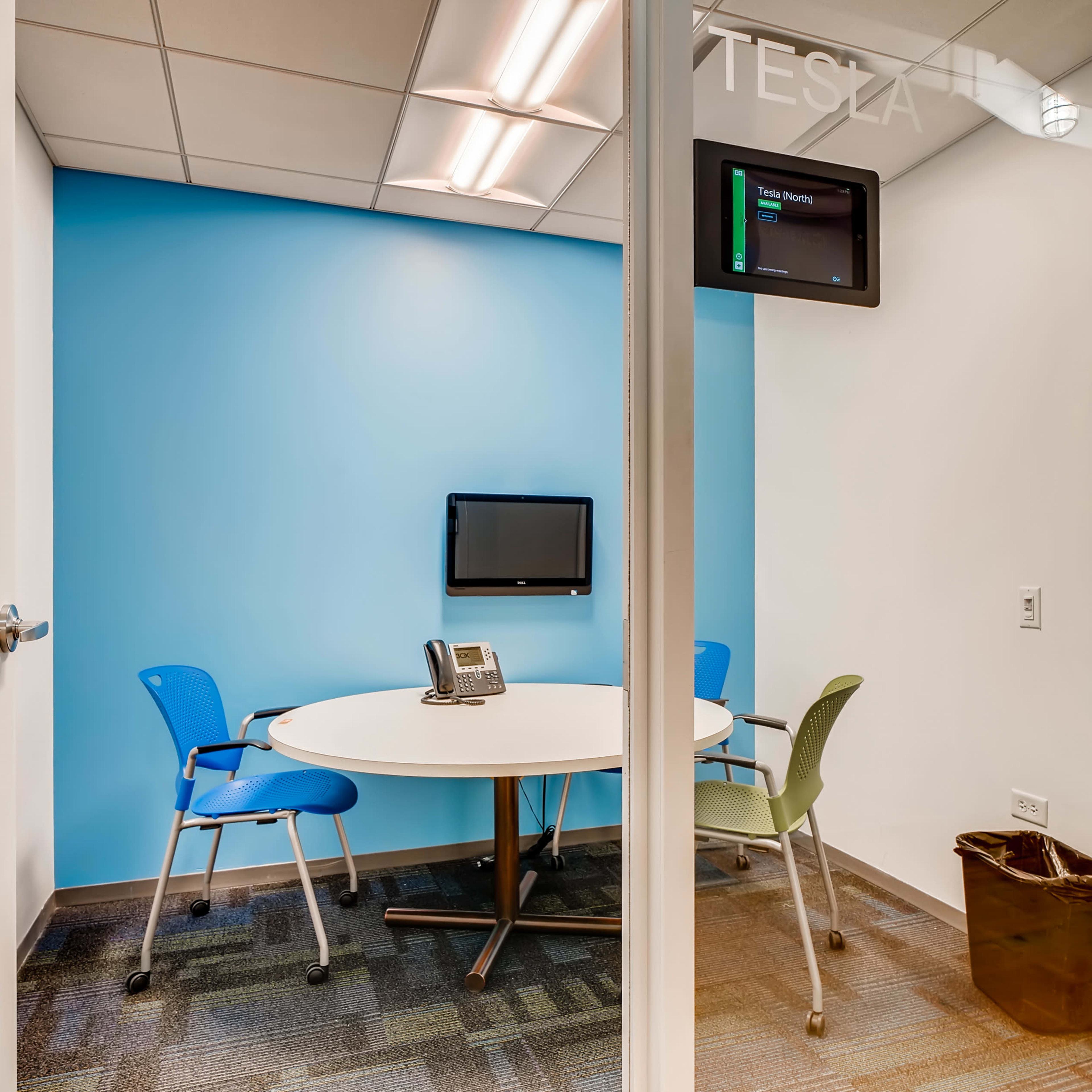 A small conference room with a round table, two chairs, a wall-mounted monitor, and a telephone, featuring a blue accent wall.
