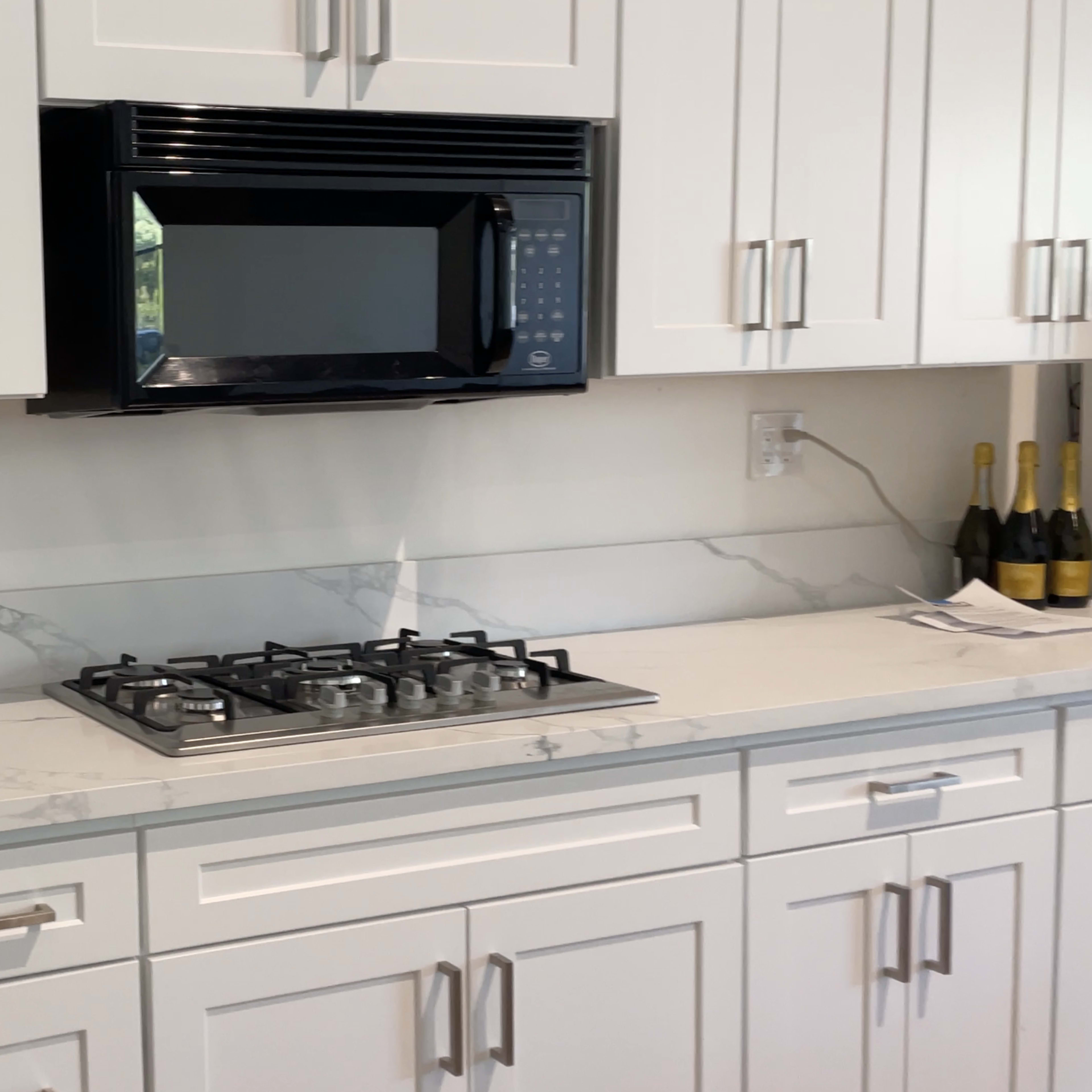 A modern kitchen features white cabinetry, a black microwave above a gas stove, and a stainless steel sink on a white marble countertop.