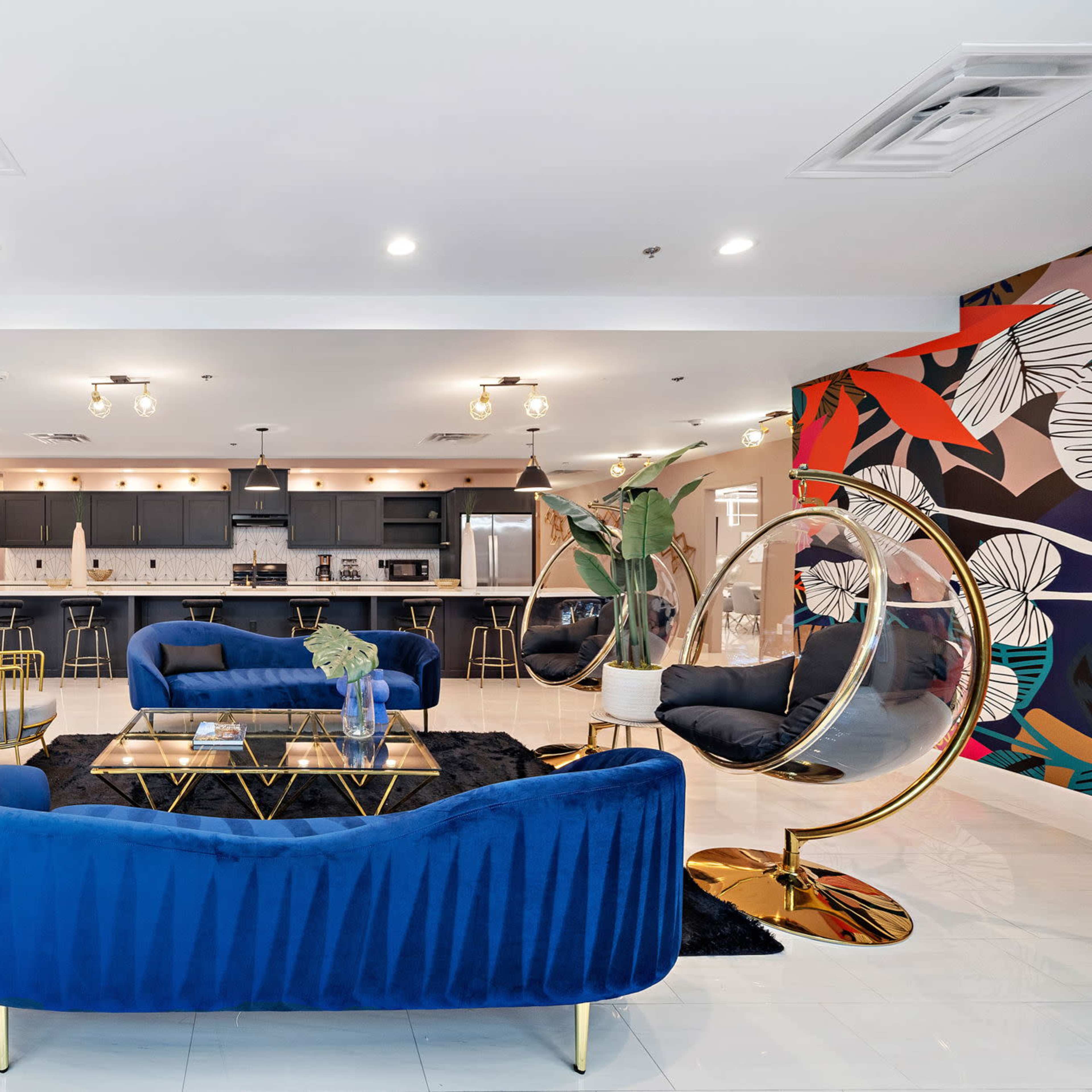 A modern lounge area with blue velvet seating, gold accents, and a colorful accent wall featuring floral designs, leading to a kitchen space in the background.