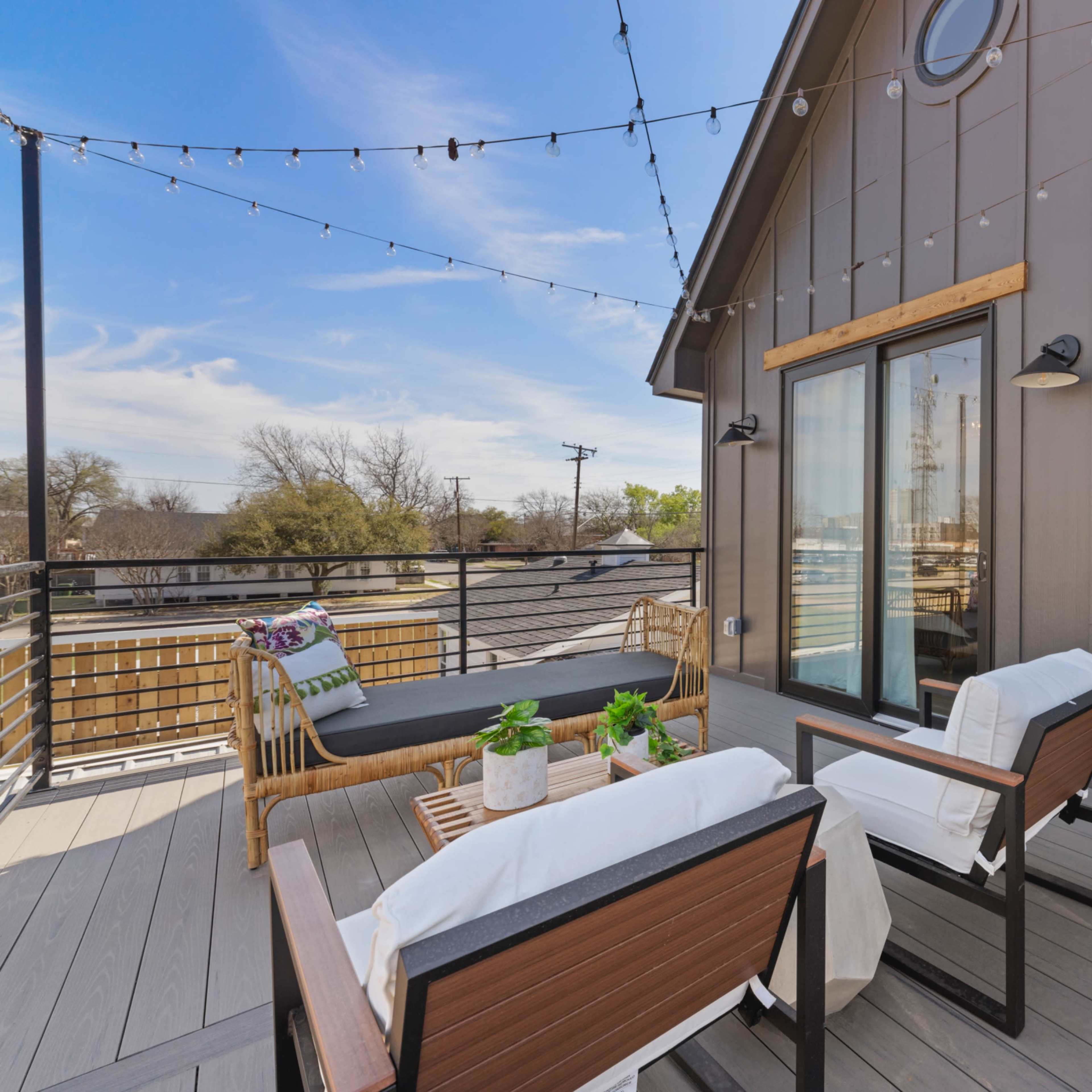 A modern outdoor terrace features a seating area with two chairs and a table, surrounded by string lights and plants.