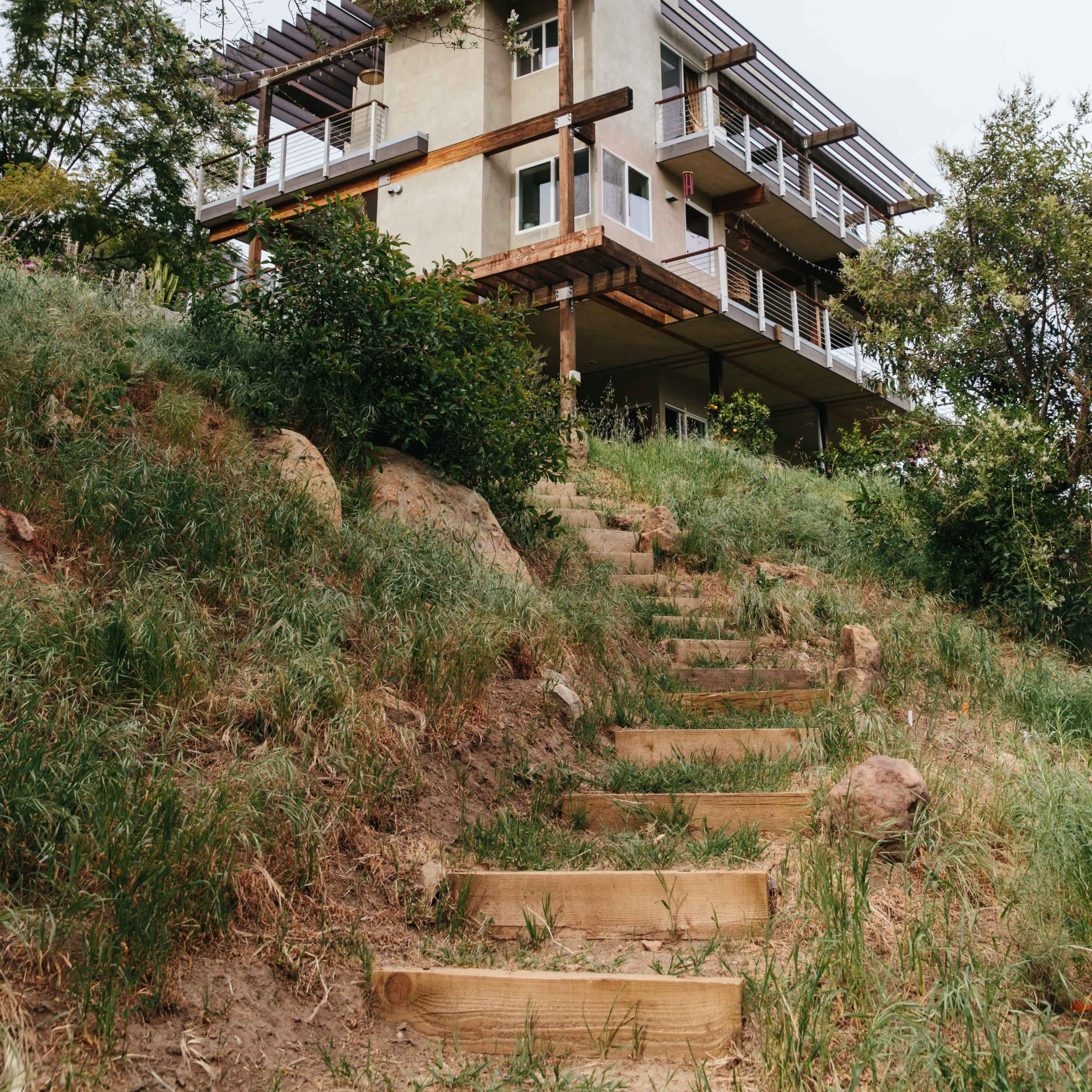 A modern house with multiple levels and a balcony is situated on a hillside, accessible by a path lined with grass and steps.