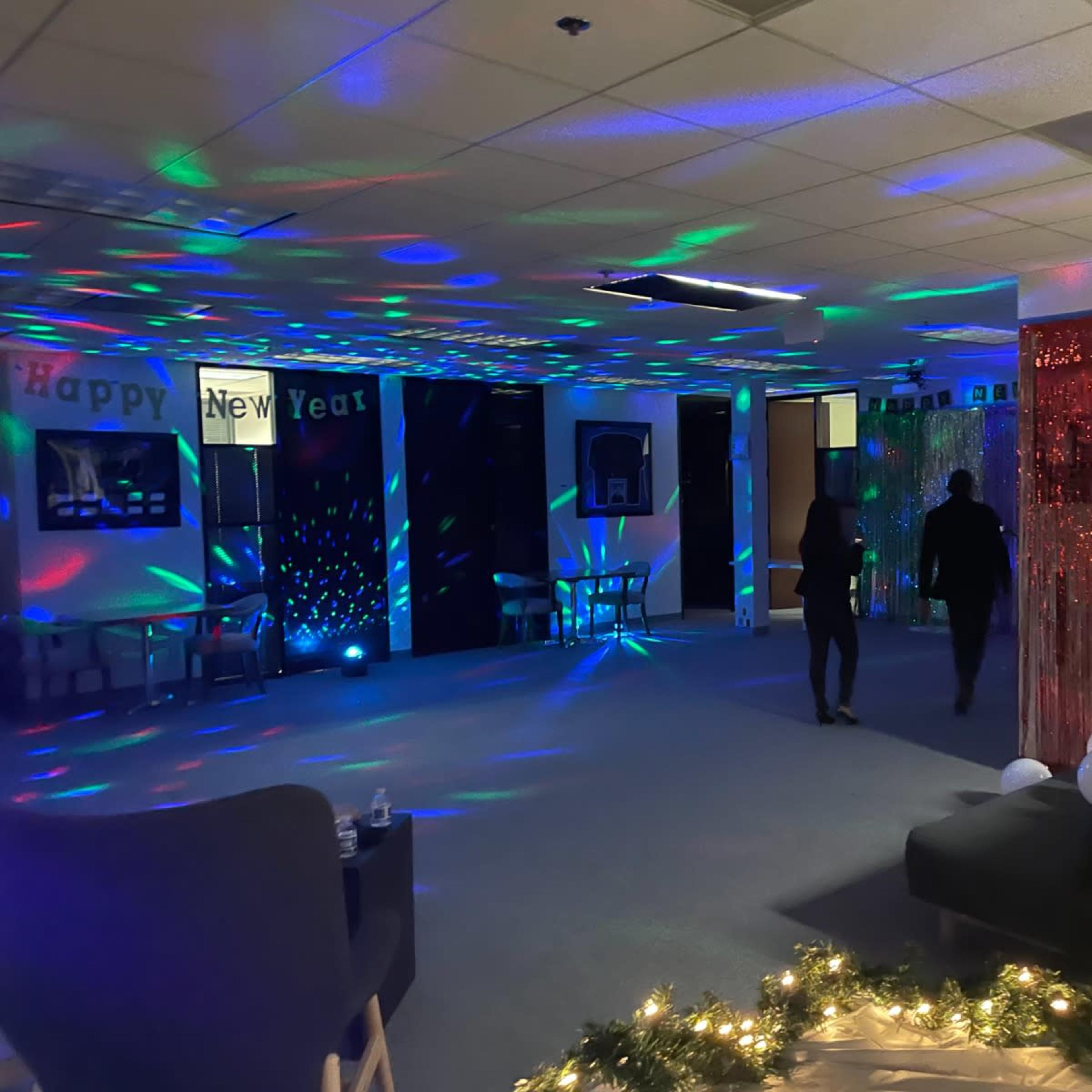 Two people walk through a decorated room with colorful lights and festive banners, preparing for a New Year's celebration.