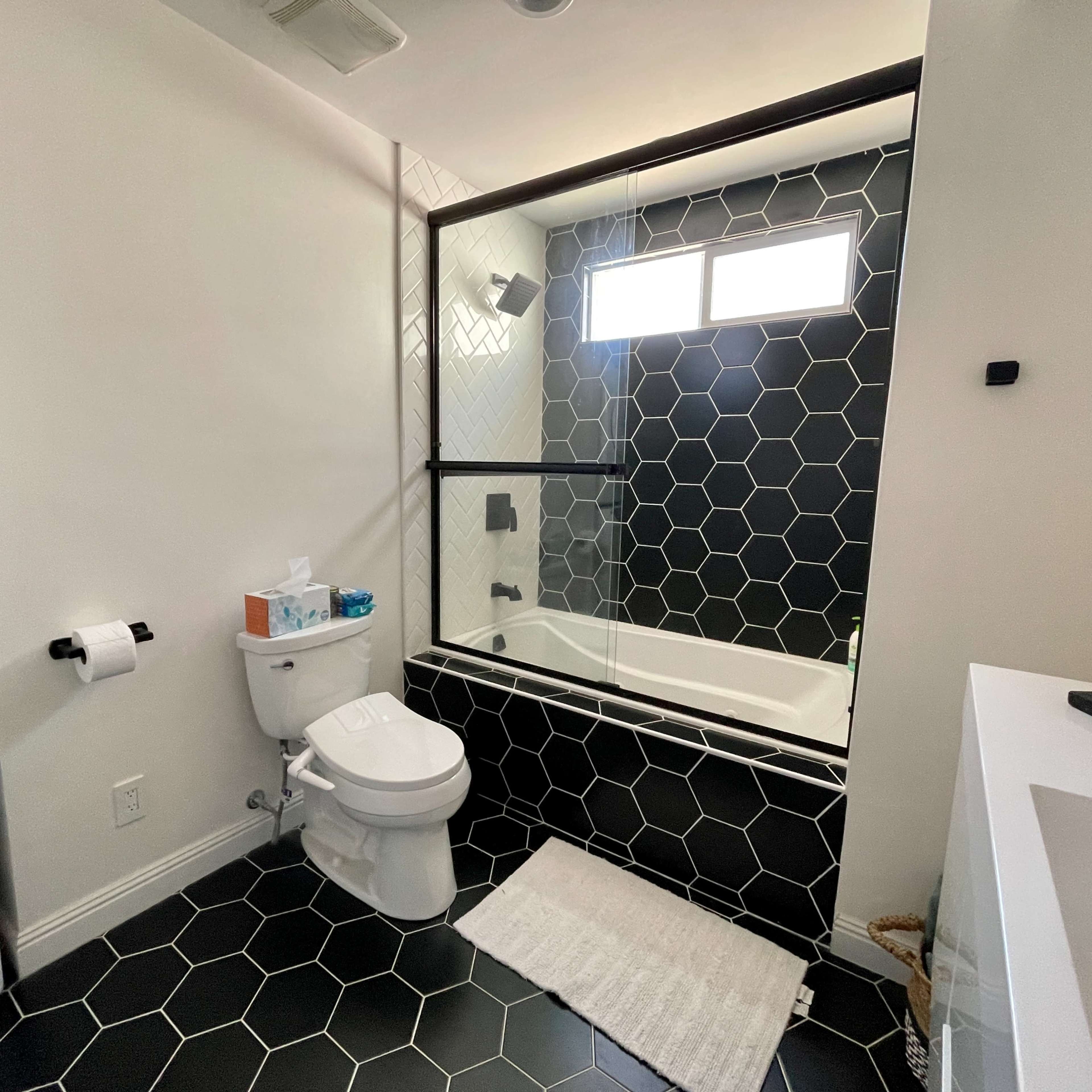 A modern bathroom with a washing machine and dryer stacked on one side, a toilet, and a shower-bathtub combination with hexagonal tile flooring.