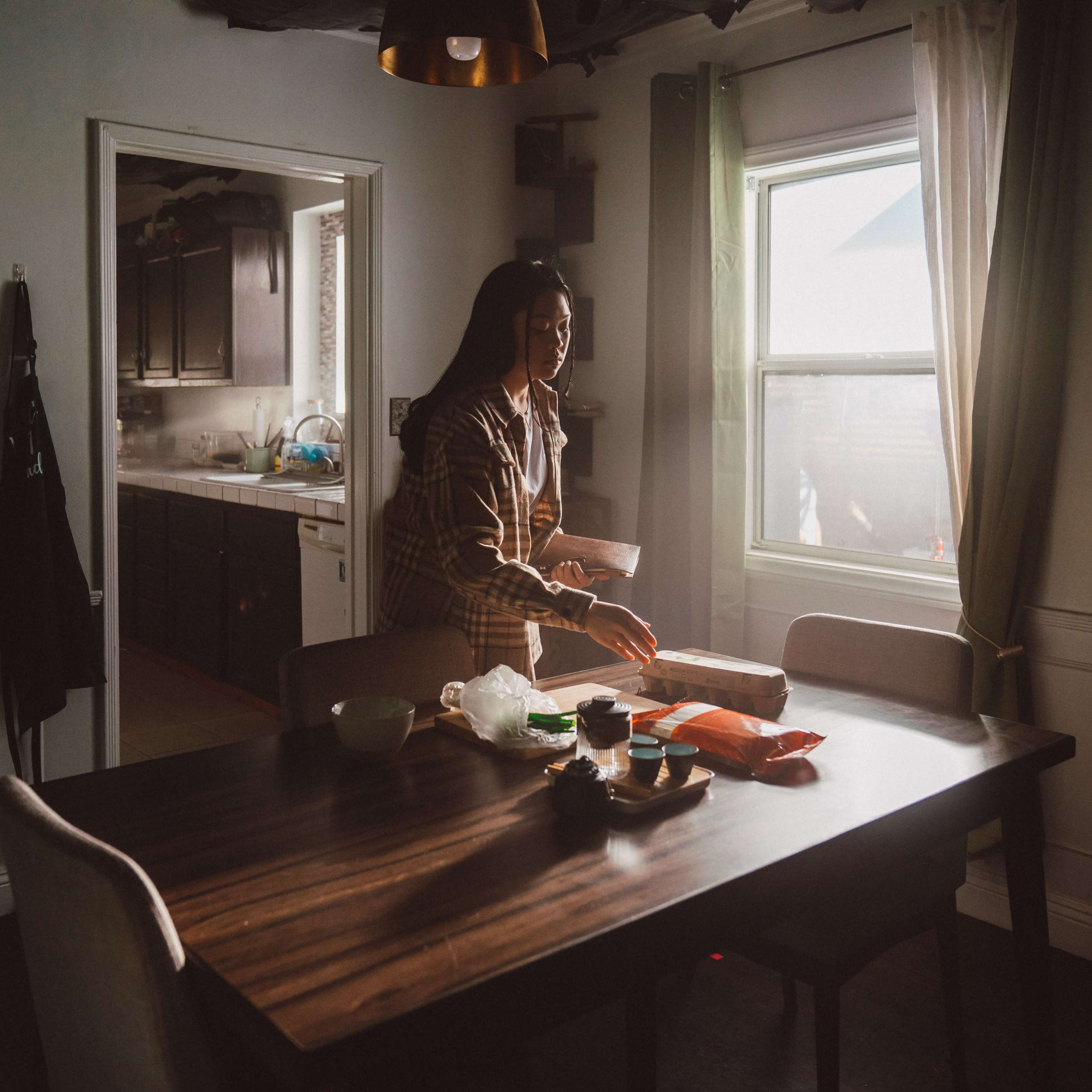 A woman stands at a dining table in a sunlit kitchen, holding a piece of paper while various food items are arranged on the table.